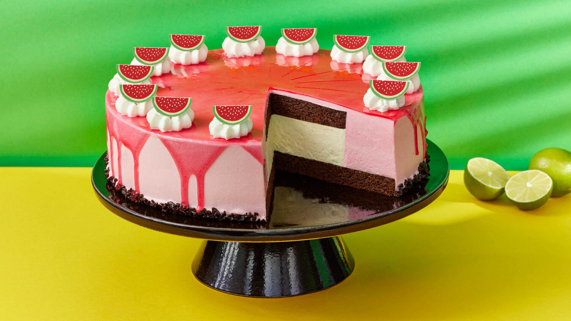 Eine rosa glasierte Torte mit Schokoladenschichten und einer Cremefüllung steht auf einem schwarzen Tortenständer. Die Torte ist mit Schlagsahne und wassermelonenförmigen Toppern dekoriert. Ein Stück fehlt, und im Hintergrund sind Limetten auf einem gelben Untergrund zu sehen.