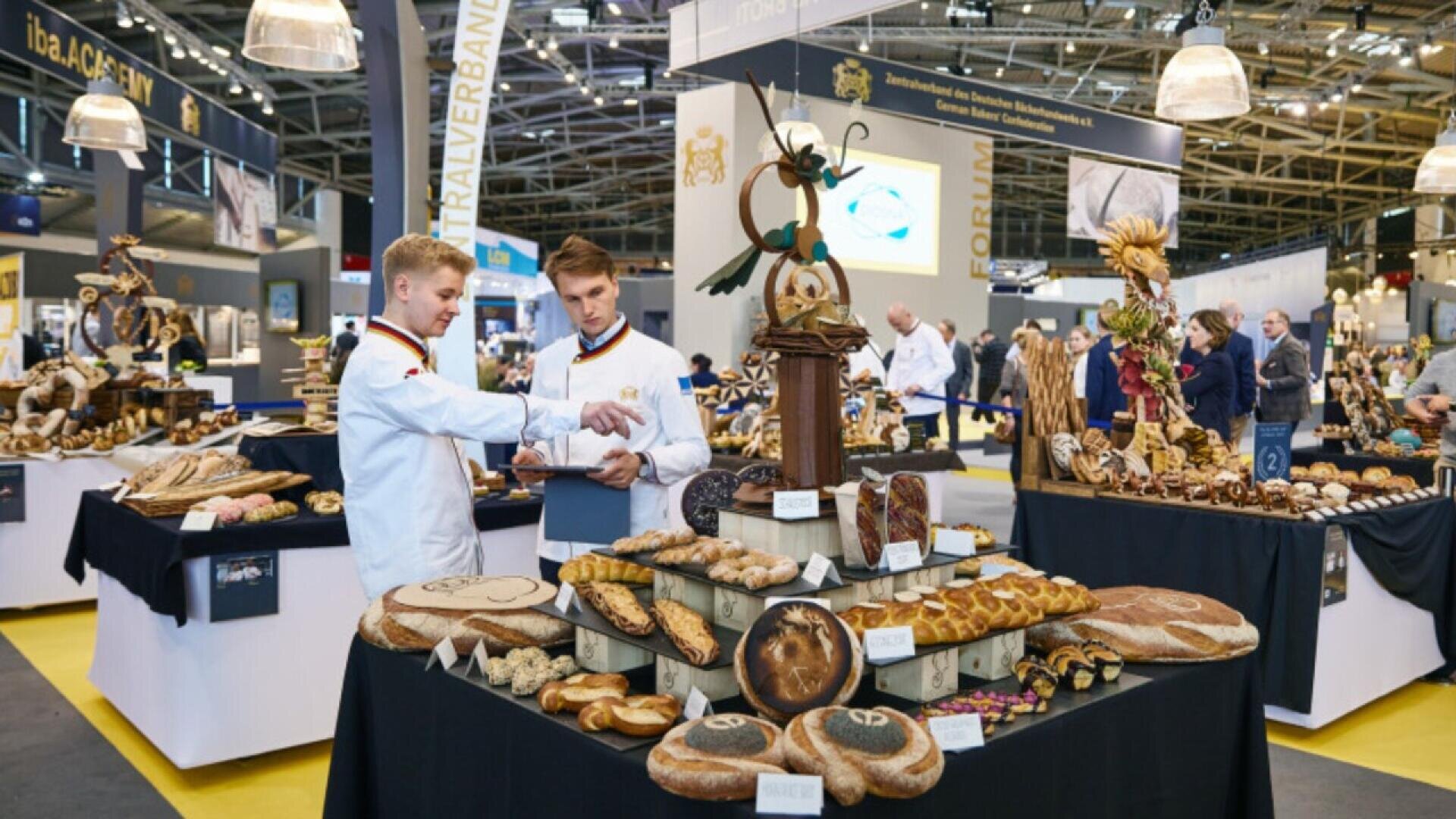 Two bakers in white uniforms stand by a table filled with assorted breads and pastries at a bakery exhibition, with more decorated tables and people in the background under bright indoor lighting.