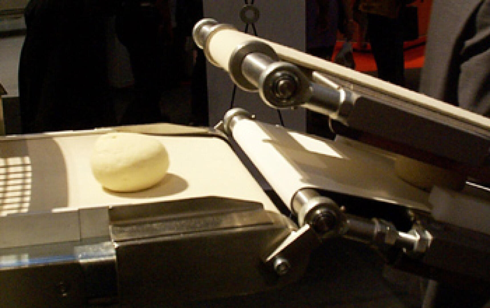 Eine Teigkugel befindet sich auf einem Förderbandsystem in einer industriellen Umgebung, das wahrscheinlich Teil einer Lebensmittelverarbeitungs- oder Bäckereimaschine ist.