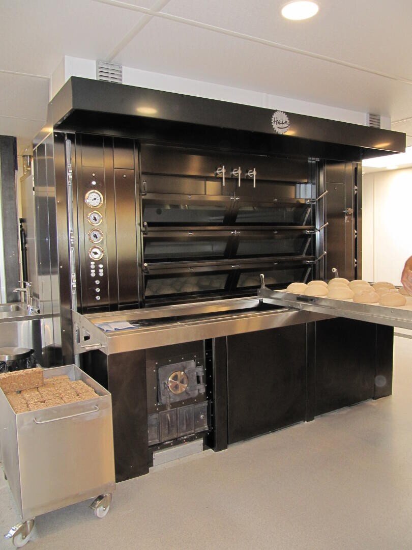 In einer Bäckerei steht ein handelsüblicher Brotofen mit mehreren Einschüben und Drehreglern. Auf einem Metalltisch davor liegen mehrere ungebackene runde Brote, und in der Nähe steht ein Wagen mit quadratischen Broten. Die Umgebung ist sauber und modern.