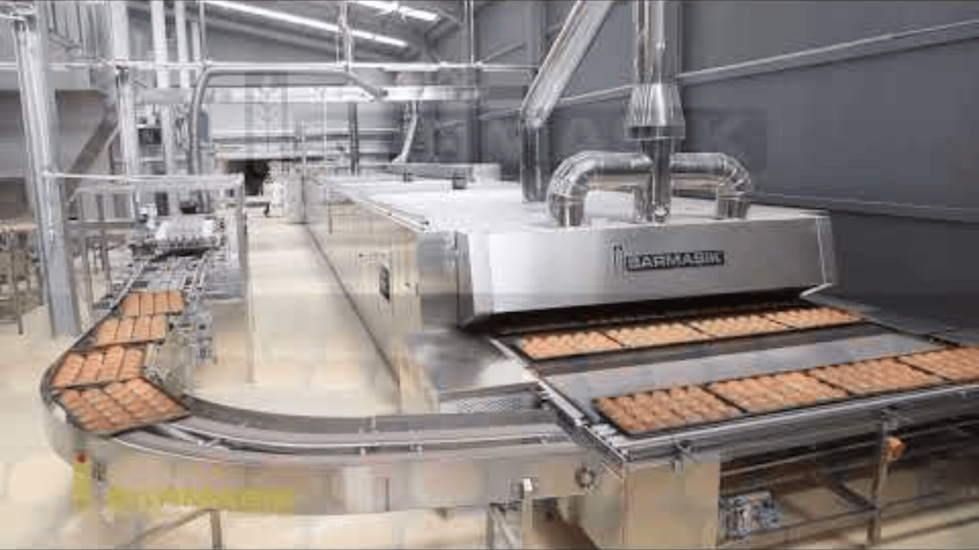 A modern industrial bakery with an automated conveyor system transports trays of baked goods through large stainless steel machines in a clean, well-lit facility.