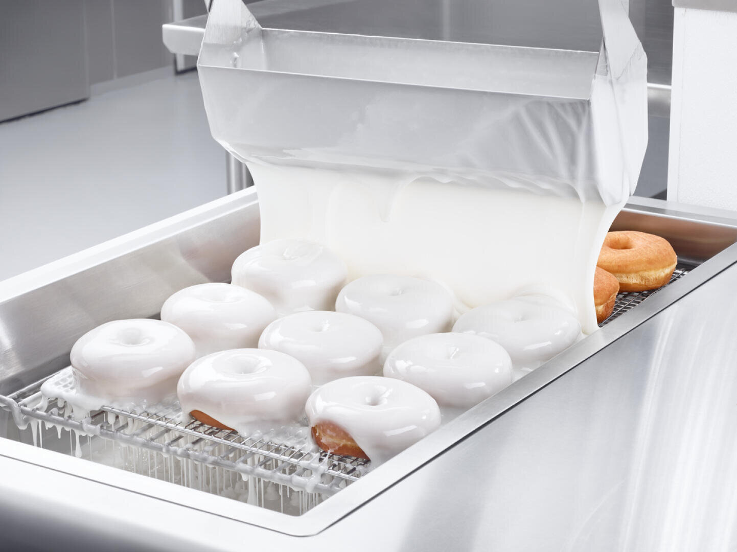 Rows of donuts on a metal rack are being covered in a thick layer of white glaze from a machine in a commercial kitchen setting.