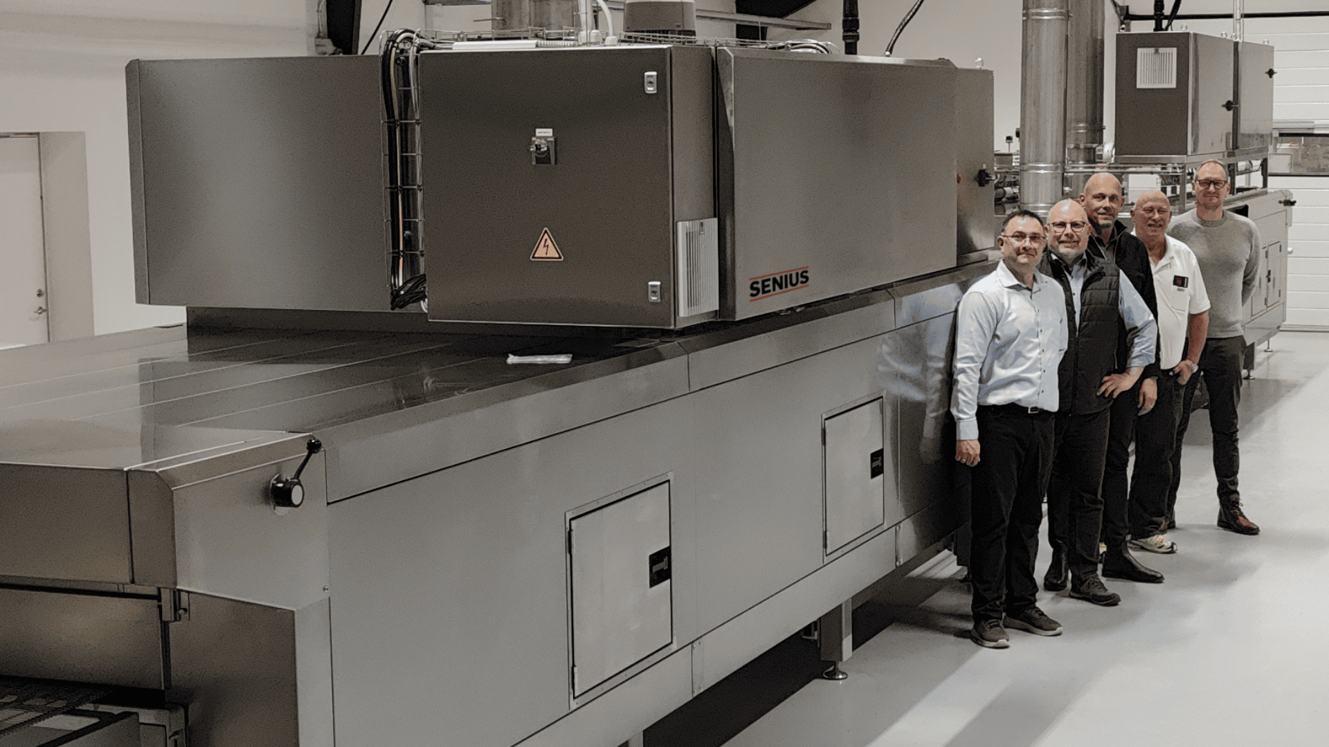 Vier Männer stehen in einer sauberen, modernen Einrichtung neben einer großen Industriemaschine aus rostfreiem Stahl mit der Aufschrift SENIUS. Die Maschine ist mit Lüftungsöffnungen, Verkleidungen und Warnschildern versehen.