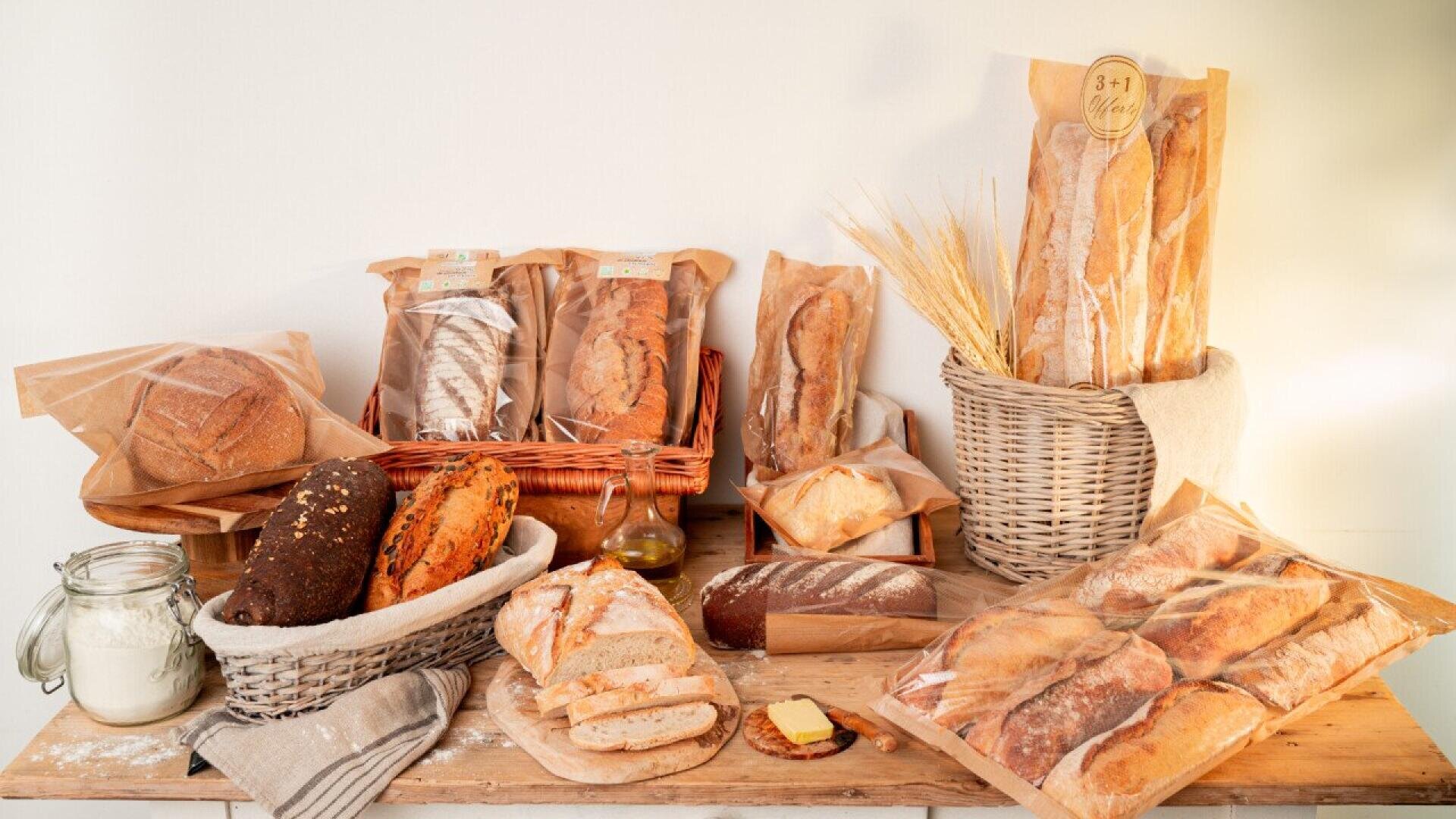 Auf einem rustikalen Holztisch liegen verschiedene Brote, Baguettes und Brötchen, teilweise in Scheiben geschnitten, in Körben und Papiertüten, daneben ein Glas Mehl, eine Schale mit Butter, ein Bündel Weizen und eine Leinenserviette.