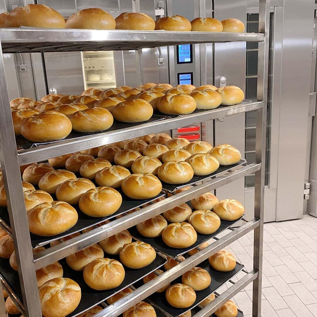 Ein Metallgestell hält mehrere Tabletts mit runden, goldbraunen Brötchen in einer gewerblichen Bäckerei mit gefliestem Boden und Edelstahlöfen im Hintergrund.