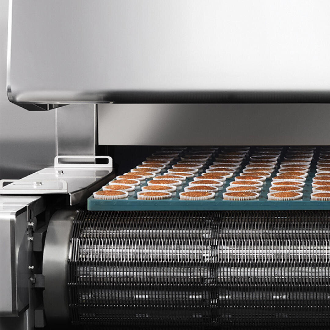 A conveyor oven baking rows of cupcake liners filled with batter, moving along a metal mesh belt inside an industrial stainless steel machine.