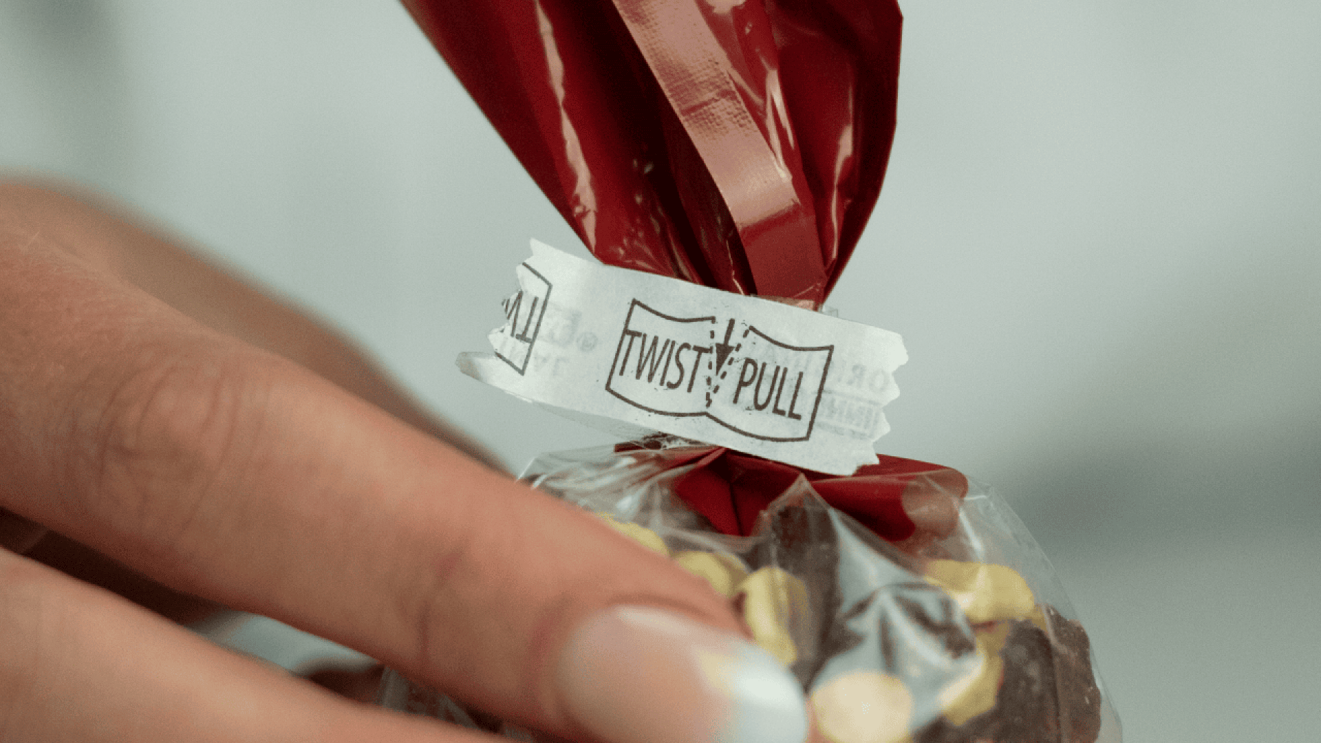 Eine Hand hält eine Plastiktüte, die mit einem roten Drehverschluss verschlossen ist. Das Etikett auf dem Band lautet TWIST & PULL. Die Tüte enthält Schokoladenbonbons, die mit gelben Bonbonstücken bestückt sind.
