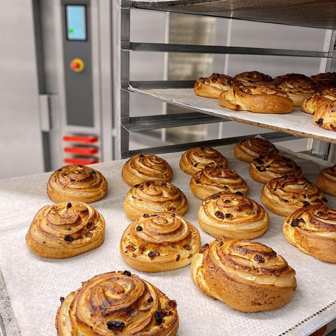 Frisch gebackenes Gebäck mit goldbraunen Strudeln und Rosinen kühlt auf mit Pergamentpapier ausgelegten Blechen auf Gestellen in einer Großküche oder Bäckerei ab.