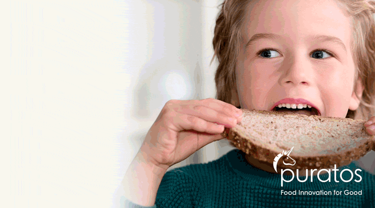 A young child in a teal sweater smiles while taking a bite from a slice of whole grain bread. The Puratos logo and slogan Food Innovation for Good appear in the bottom right corner.