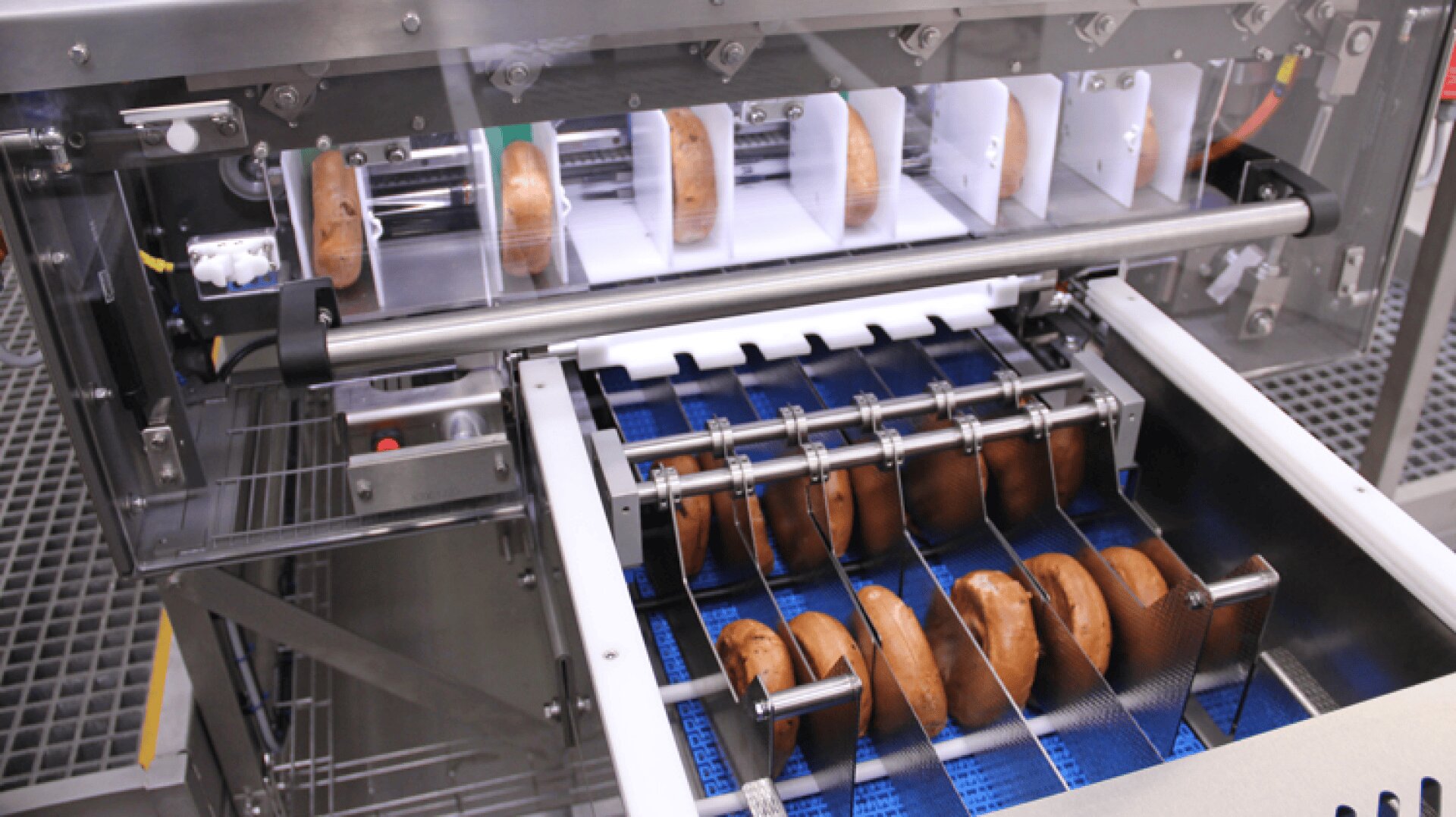 A machine slicing bagels on a conveyor belt in a factory setting. The bagels move through a series of blades that cut them in half, with blue and metal parts visible in the equipment.