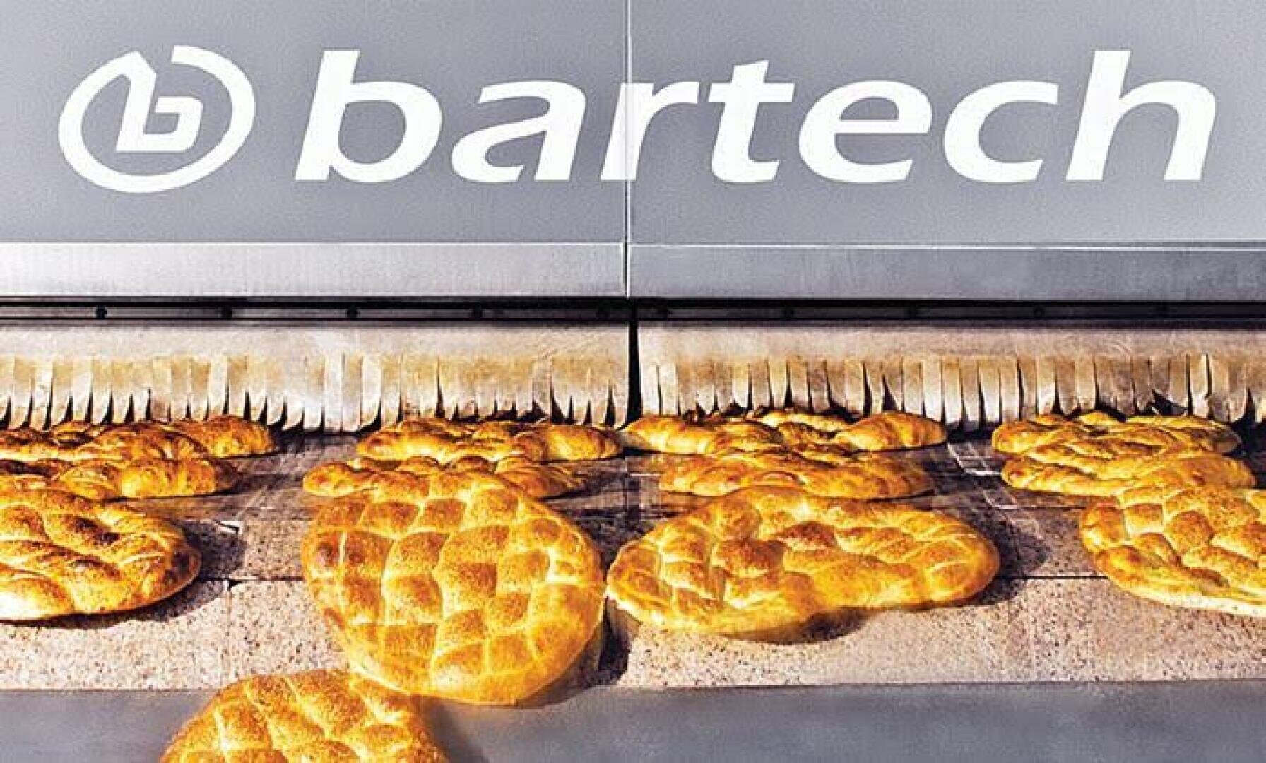 In a bakery or industrial bakery, freshly baked loaves of bread run along a conveyor belt under the large bartech company logo.