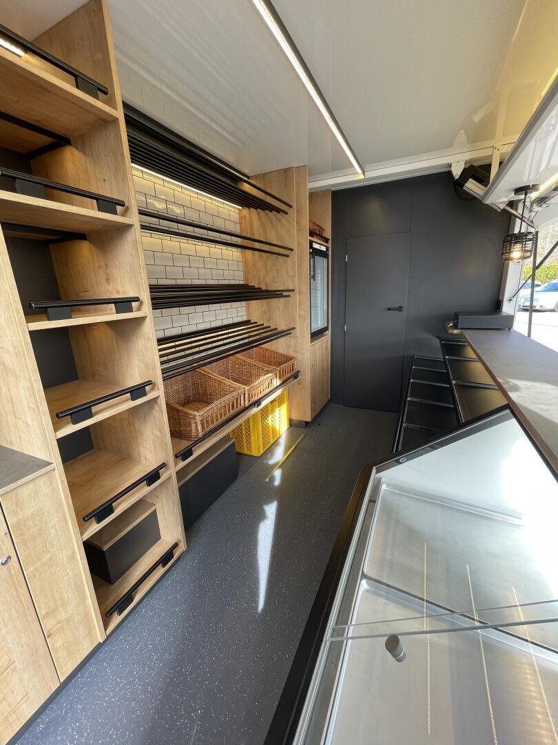 Innenraum eines modernen Foodtrucks oder mobilen Ladens mit leeren Holzregalen, Weidenkörben, einer schwarzen Theke, einer gefliesten Wand und einer Glasvitrine, alles gut beleuchtet mit natürlichem und künstlichem Licht.