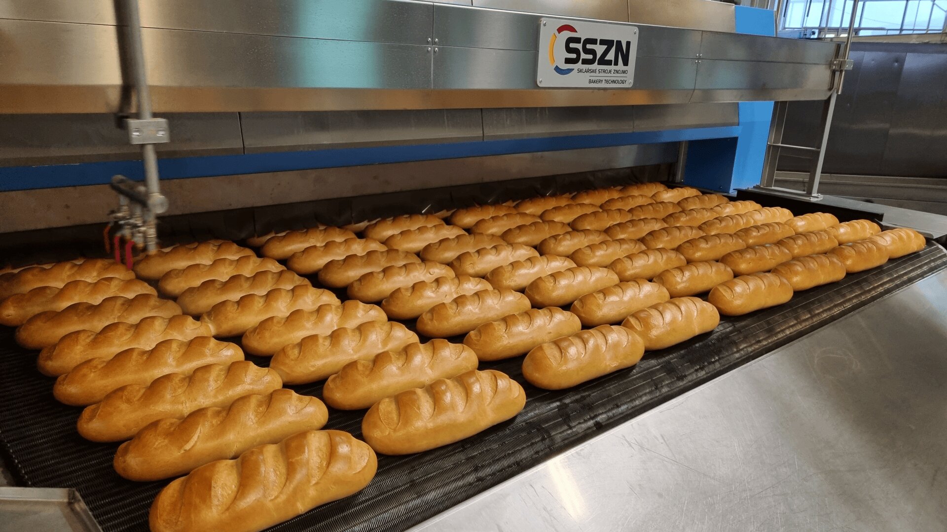 Rows of freshly baked loaves of bread on a conveyor belt in an industrial bakery, with a large stainless steel machine labeled SSZN in the background.