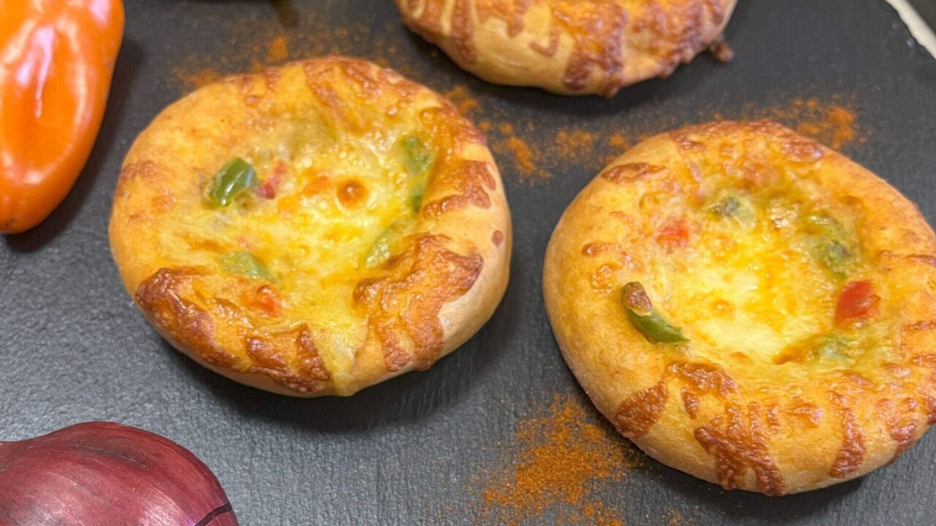 Mini cheese and vegetable tarts with golden-brown crusts on a black surface. A red onion and an orange bell pepper are partially visible beside the tarts.