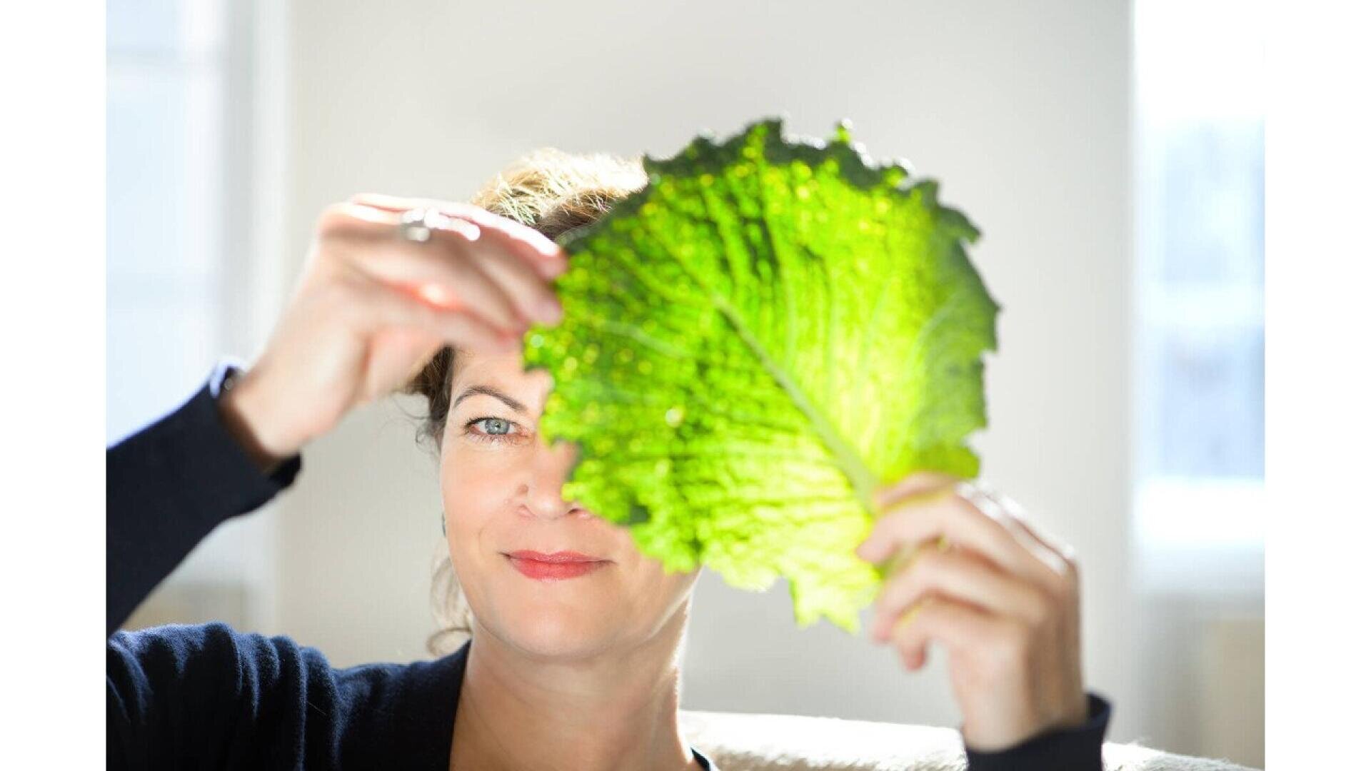 A woman holds a large, bright green leaf in front of her face, partially covering one eye while sunlight shines through the leaf, creating a vivid, translucent effect.