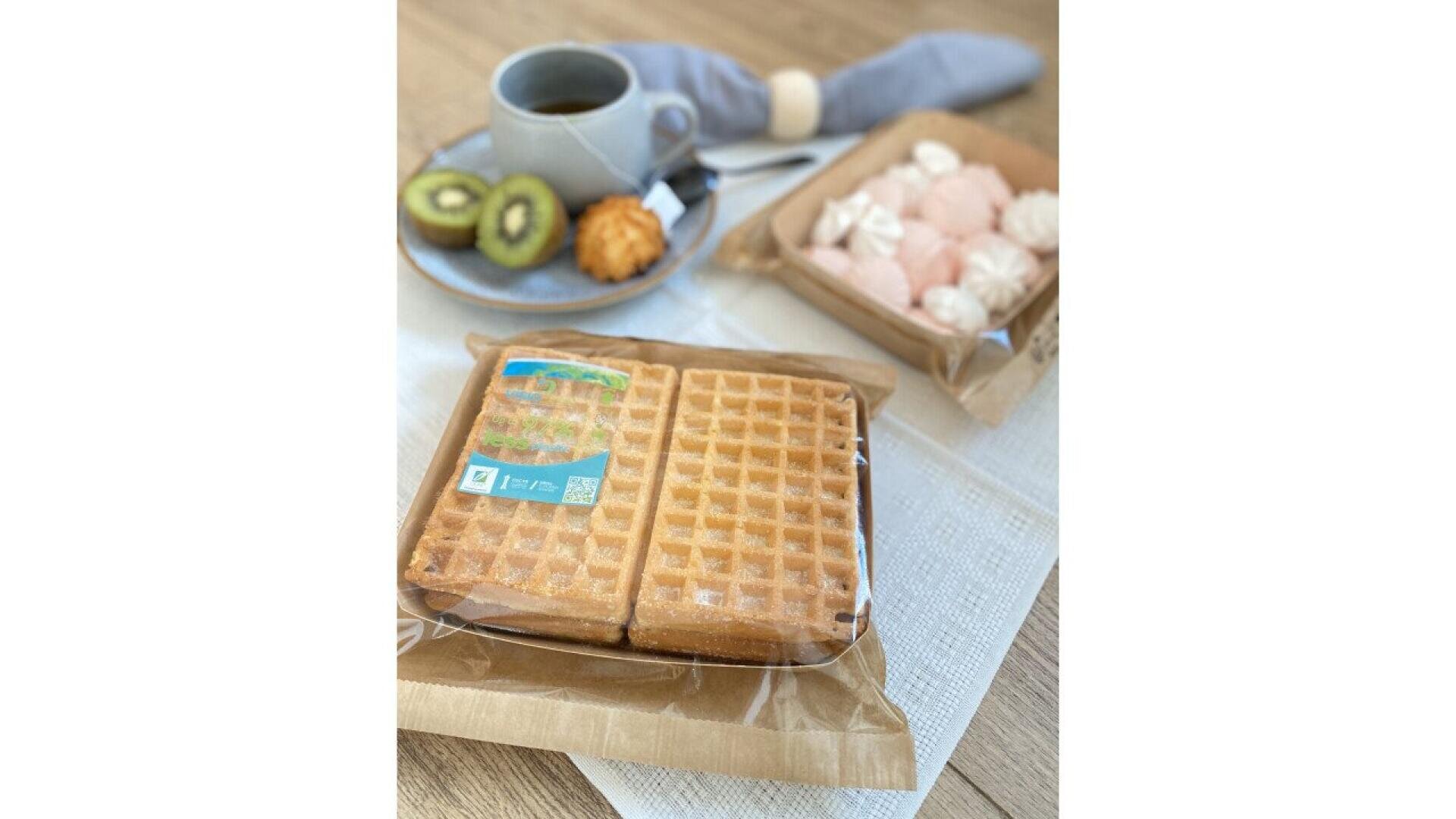 Ein Paket mit zwei Waffeln in Plastikverpackung auf einem Tisch. Im Hintergrund stehen auf einem Teller eine Tasse Kaffee, zwei Kiwischeiben, ein Gebäckstück und eine Schachtel mit rosa und weißen Baiser-Keksen.