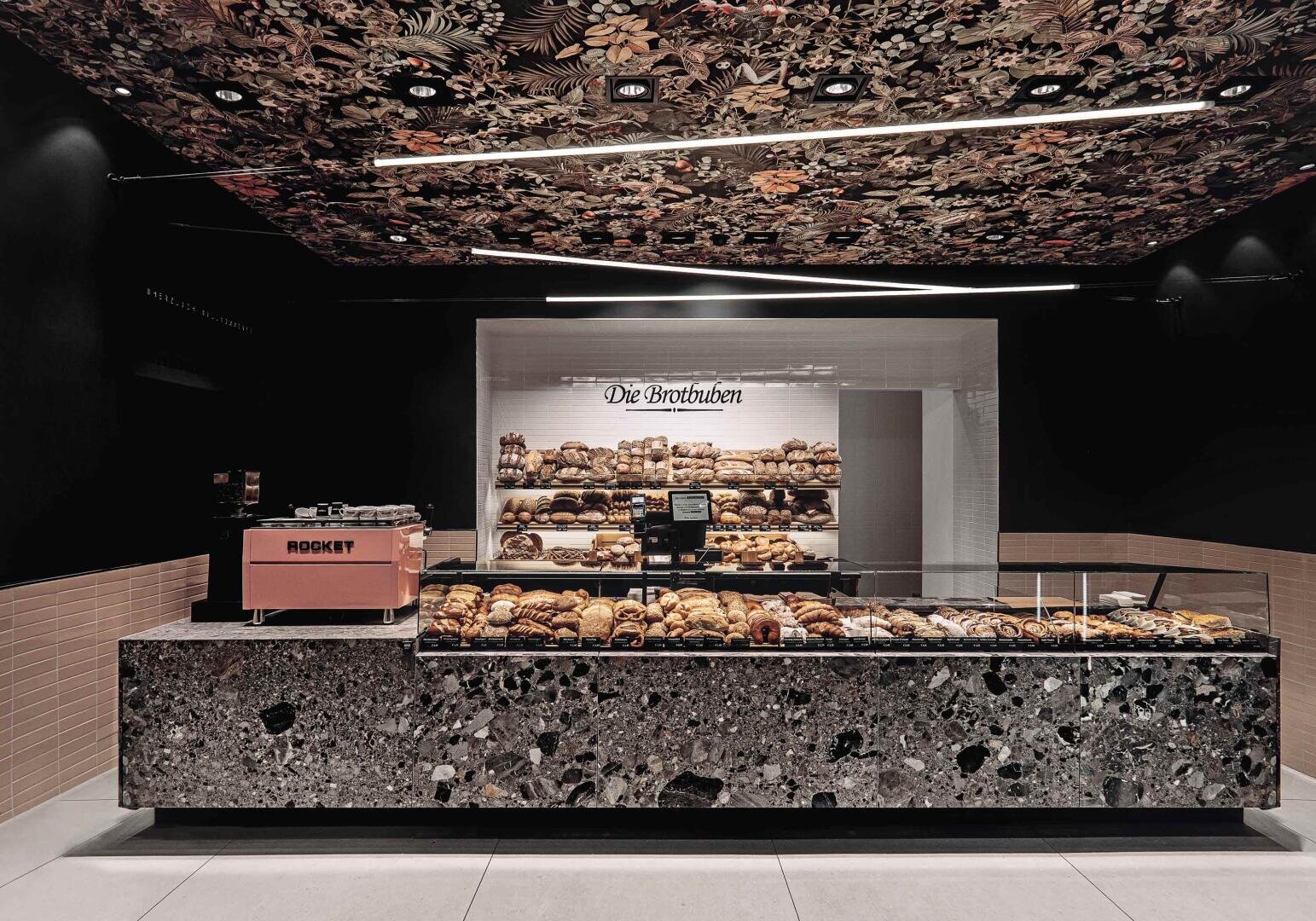 Eine moderne Bäckerei mit einer Terrazzo-Theke mit verschiedenen Backwaren, einer rosafarbenen Espressomaschine und Regalen voller Brot. Die Decke hat ein dunkles Blumenmuster und auf dem Schild über dem Brot steht "Die Brotbuben".