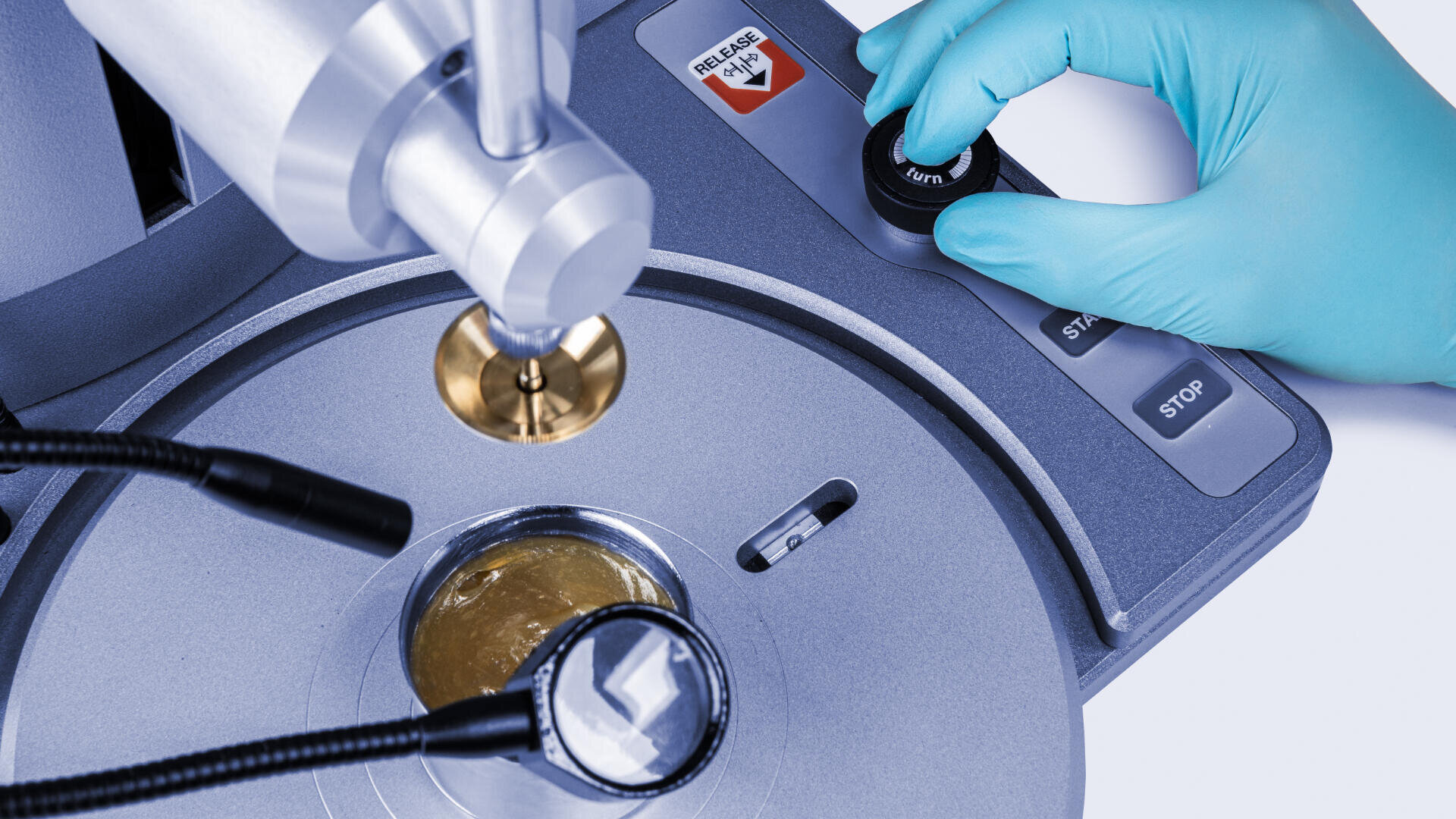 Nahaufnahme einer behandschuhten Hand, die ein Laborgerät mit Metallteilen und mit Start und Stopp beschrifteten Tasten bedient. Die Maschine scheint eine Probe zu analysieren oder zu verarbeiten.