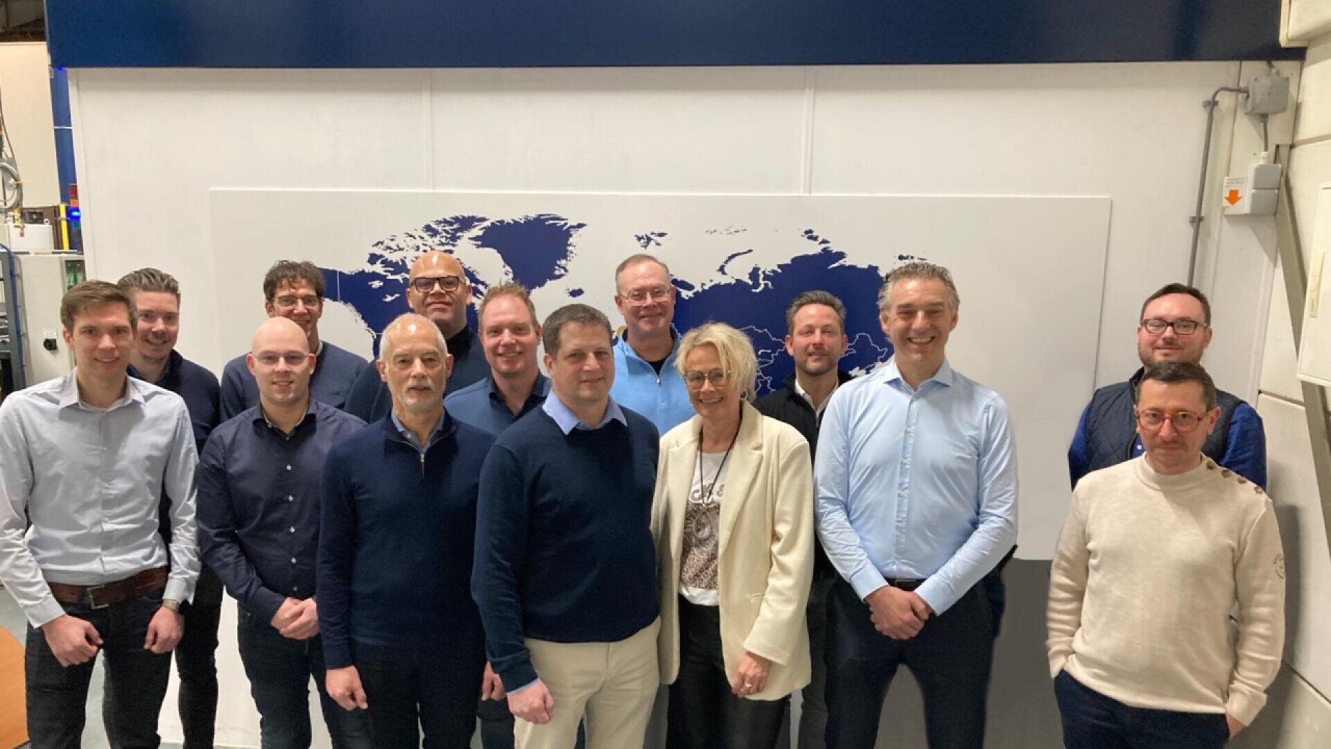 A group of thirteen people, including men and one woman, stand indoors in front of a world map, posing and smiling for a group photo. Most are dressed in business or business casual attire.