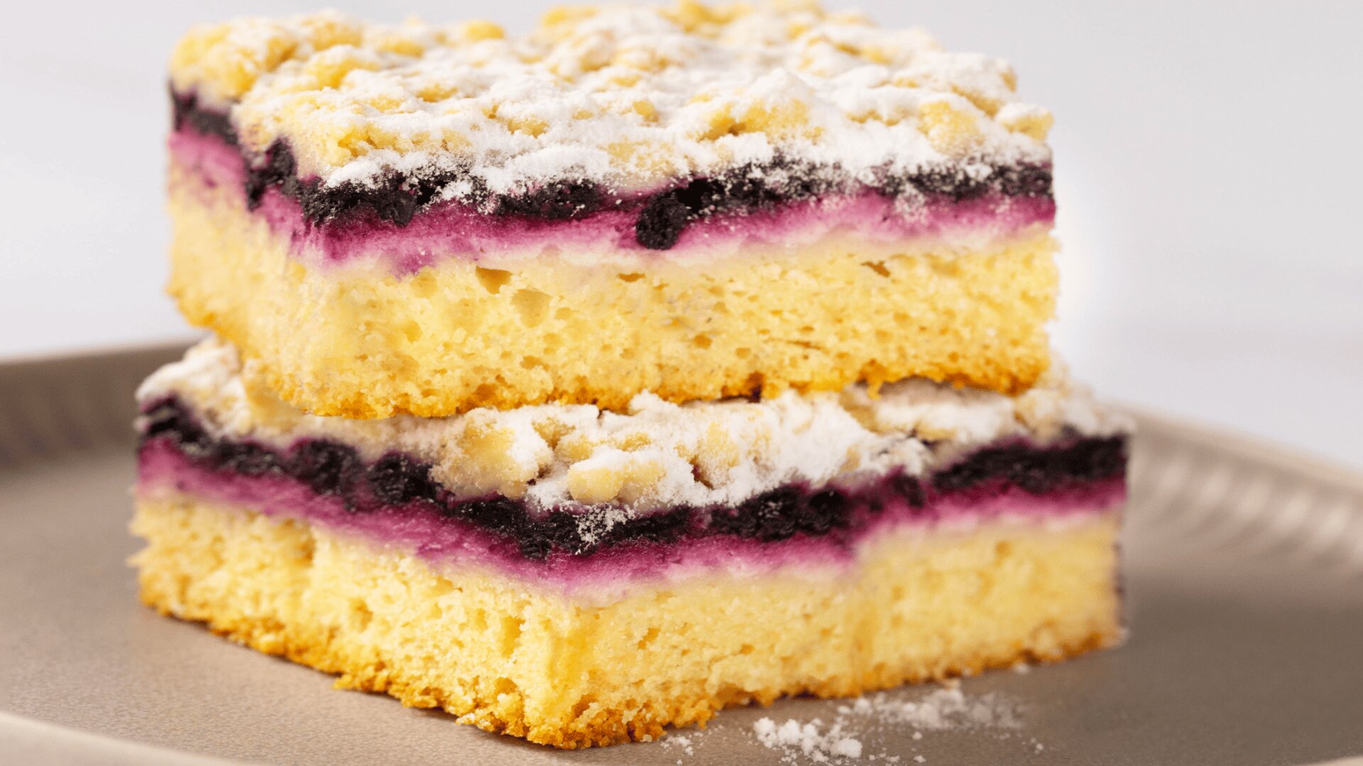 Zwei quadratische Scheiben Streuselkuchen mit gelbem Boden, lilafarbener Beerenfüllung und mit Puderzucker bestäubtem Streuselkuchen, gestapelt auf einem beigen Teller.