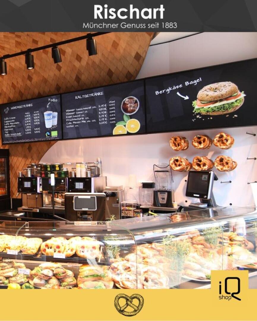 A bakery counter at Rischart in Munich displays sandwiches, pastries, and pretzels, with a menu overhead and pretzels hanging on the wall. The setting is bright and inviting, featuring modern decor.