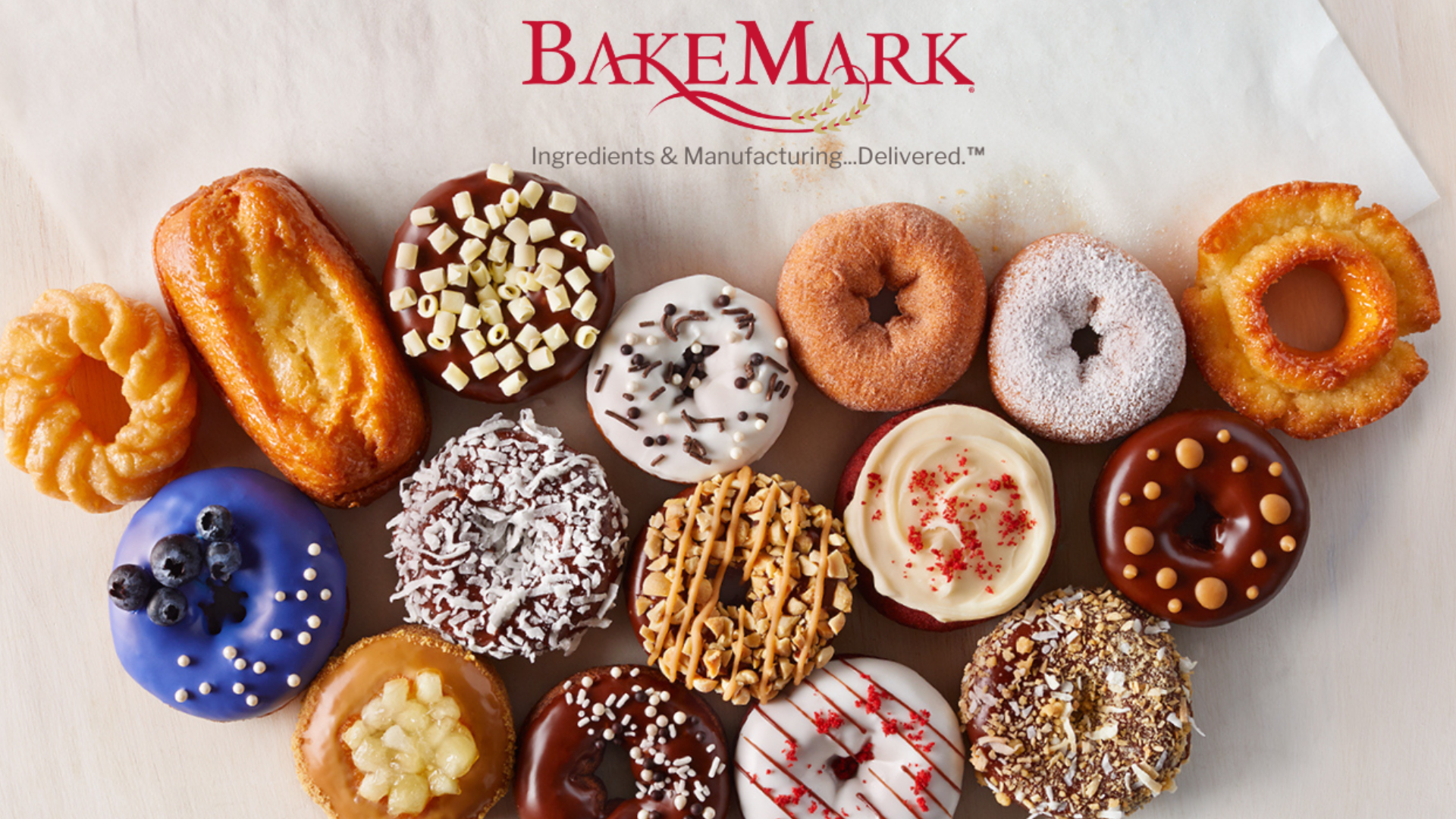 A selection of fifteen assorted donuts with various colorful toppings and glazes is arranged in a semi-circle beneath the BakeMark logo on a white background.