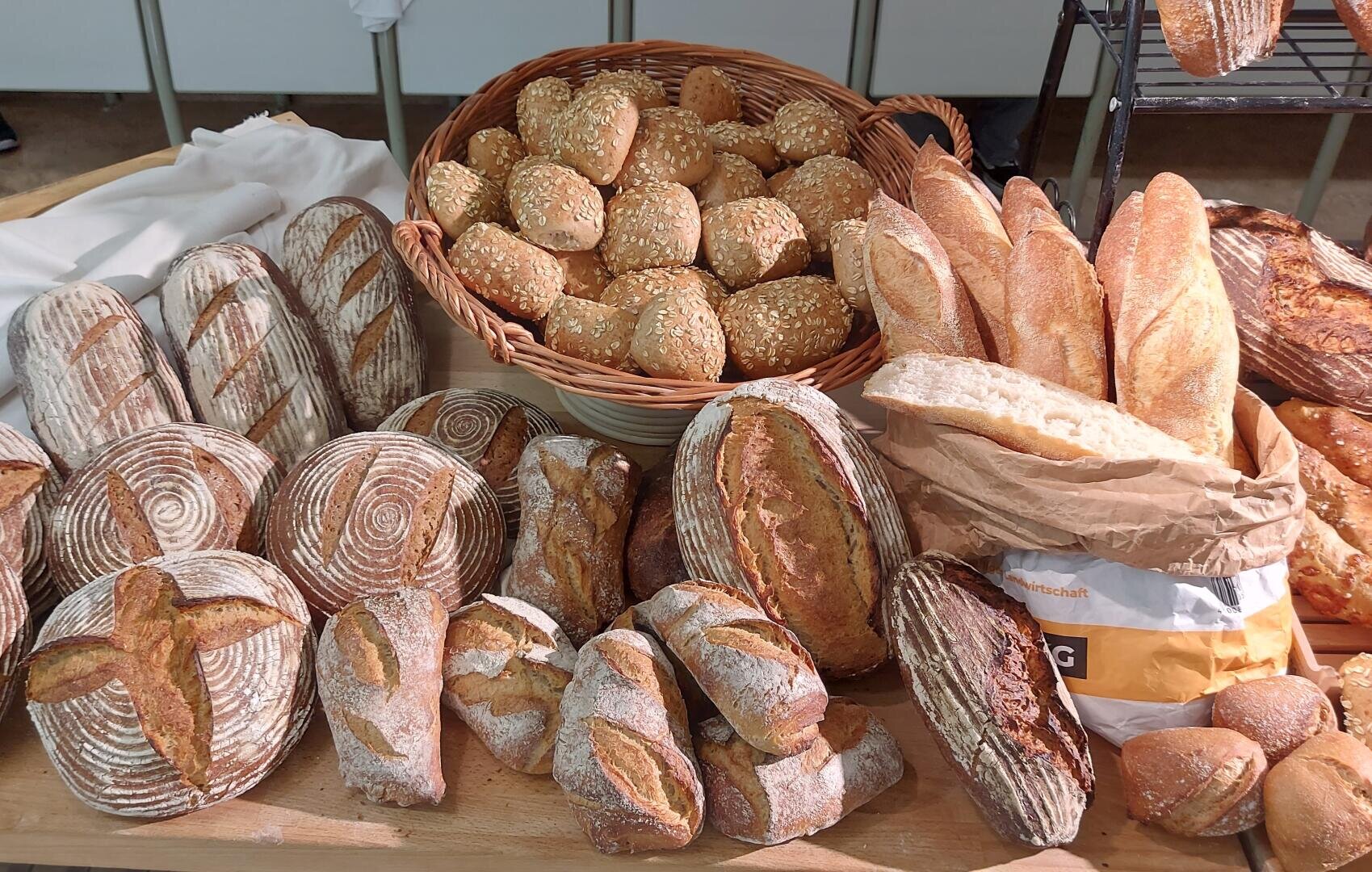 Auf einem Holztisch werden verschiedene handwerklich hergestellte Brote präsentiert, darunter runde Laibe mit dekorativen Einschnitten, Baguettes in einer Papiertüte und Sesambrötchen in einem Weidenkorb.
