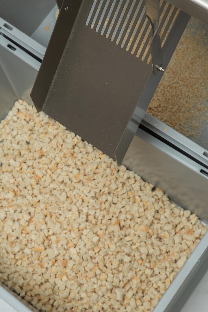 Eine Metallrutsche gibt kleine, gleichmäßig geschnittene Brotwürfel in einen großen grauen Behälter aus, der wahrscheinlich in der industriellen Lebensmittelverarbeitung oder Bäckerei verwendet wird.