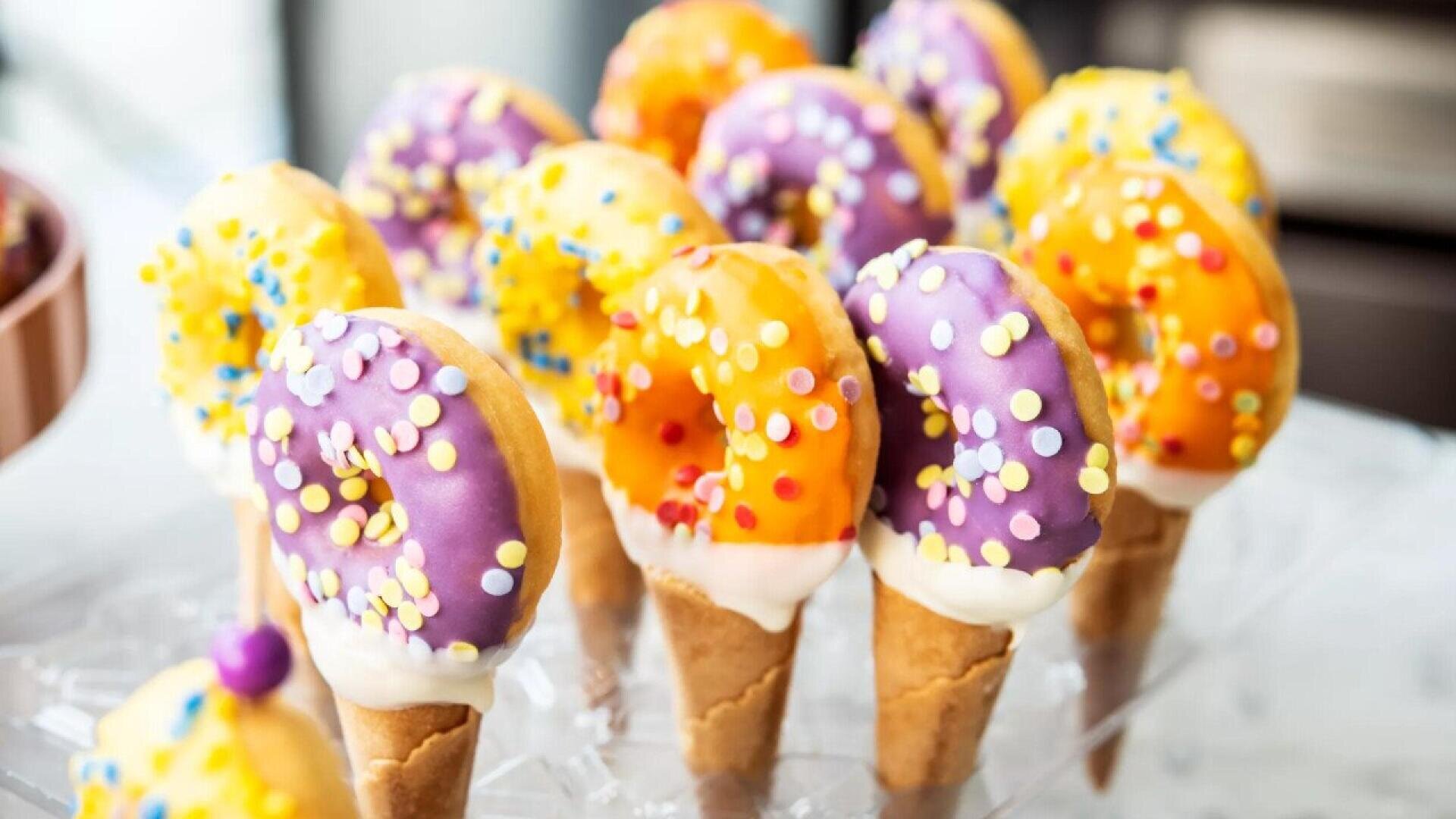 Colorful donuts with sprinkles are served on top of ice cream cones, creating a fun and whimsical dessert display. The donuts are coated in purple and orange icing and arranged upright on a clear stand.