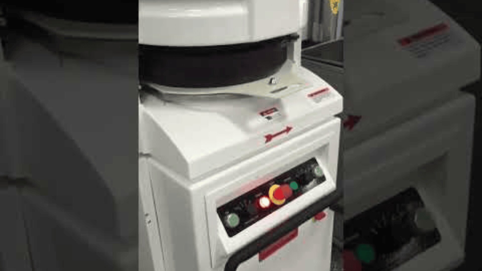A close-up of an industrial machine with a control panel featuring colored buttons and switches. The machine is primarily white with red labels and arrows, and appears to be used in a manufacturing or processing setting.