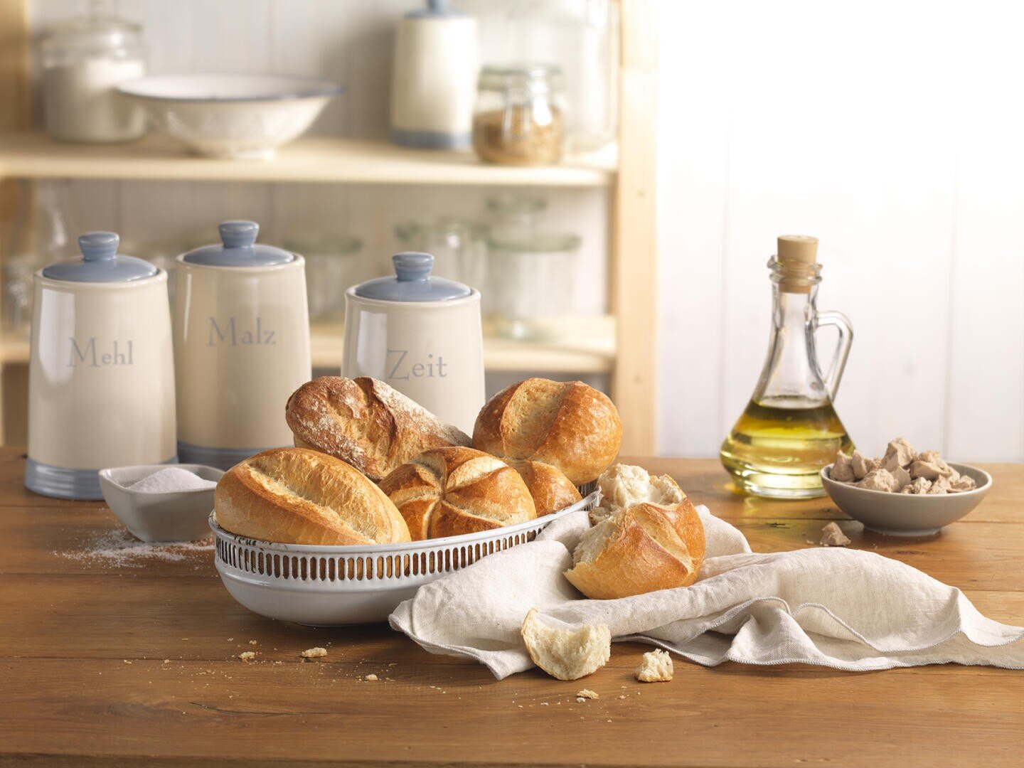 A bowl of assorted bread rolls sits on a wooden table with a linen cloth, next to a jar of olive oil, a plate of fresh yeast, and three labeled canisters in the background.