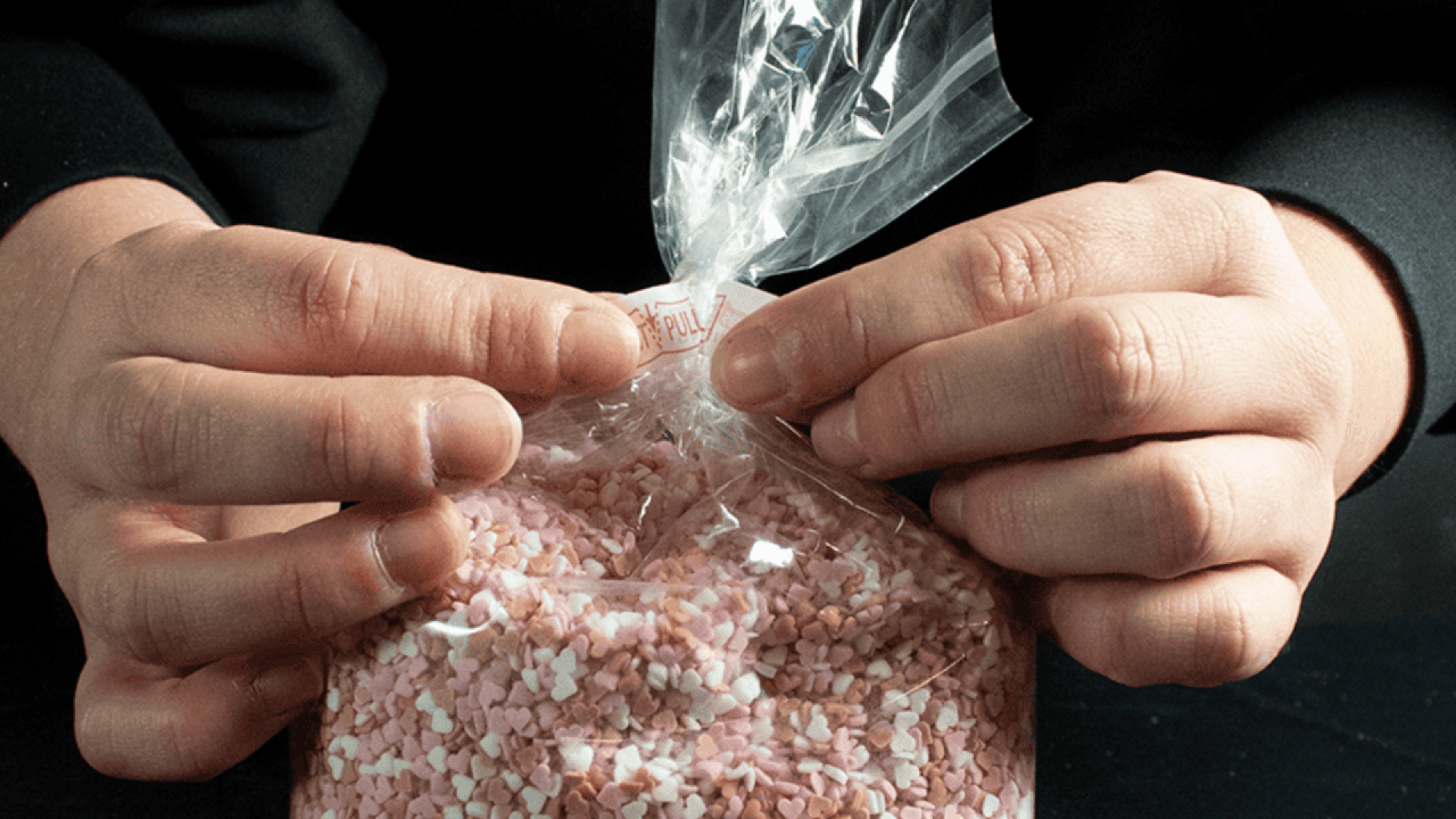 Die Hände einer Person verschließen eine durchsichtige Plastiktüte, die mit kleinen rosa und weißen herzförmigen Streuseln gefüllt ist.