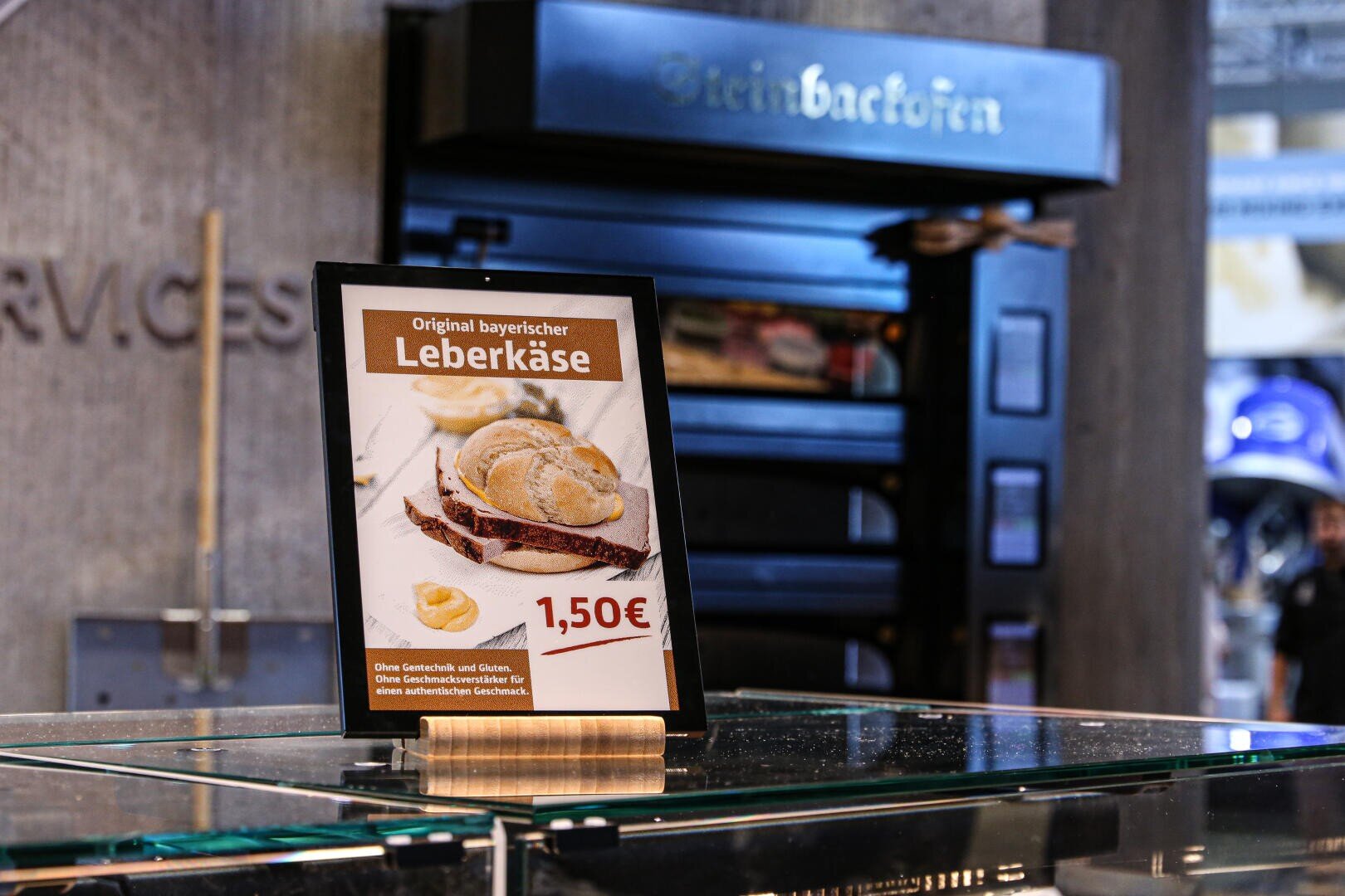 A sign displays a sandwich with Leberkäse and a bun, priced at 1,50€. The text is in German, advertising “Original bayerischer Leberkäse.” A bakery oven is visible in the background.