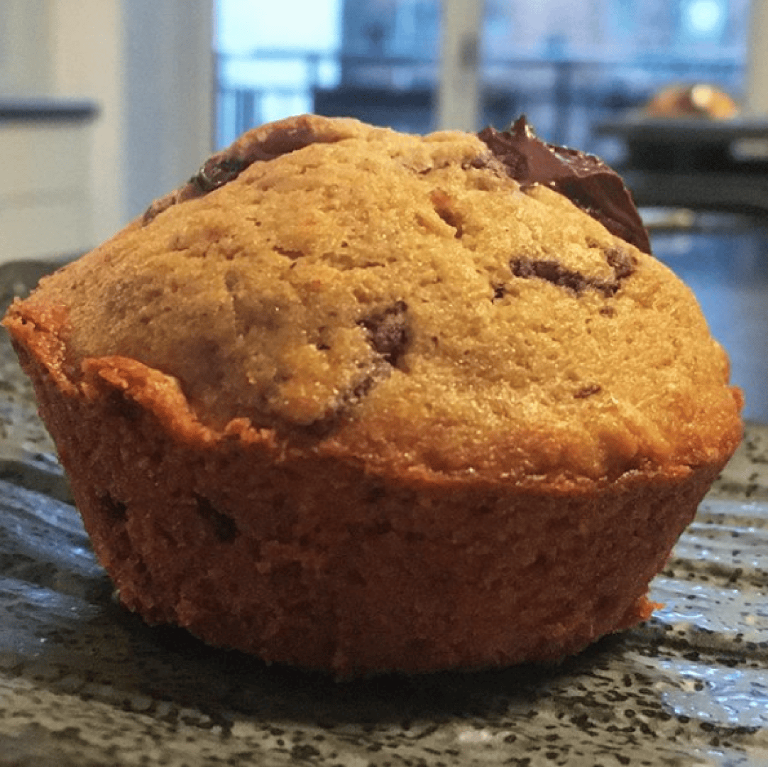A close-up of a golden-brown muffin with visible chocolate chunks, sitting on a textured dark plate. The background shows a blurred kitchen and window.