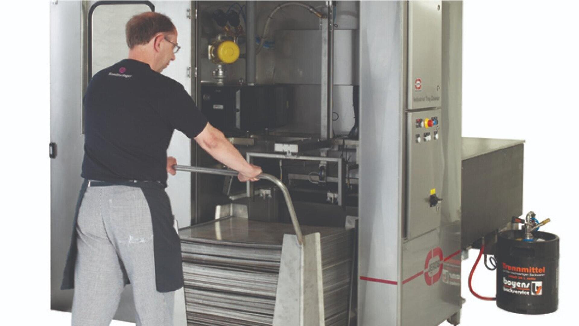 A man operates a large industrial dishwashing machine, loading trays into the machine. He is wearing glasses and an apron, and nearby there is a container labeled Fassmittel.