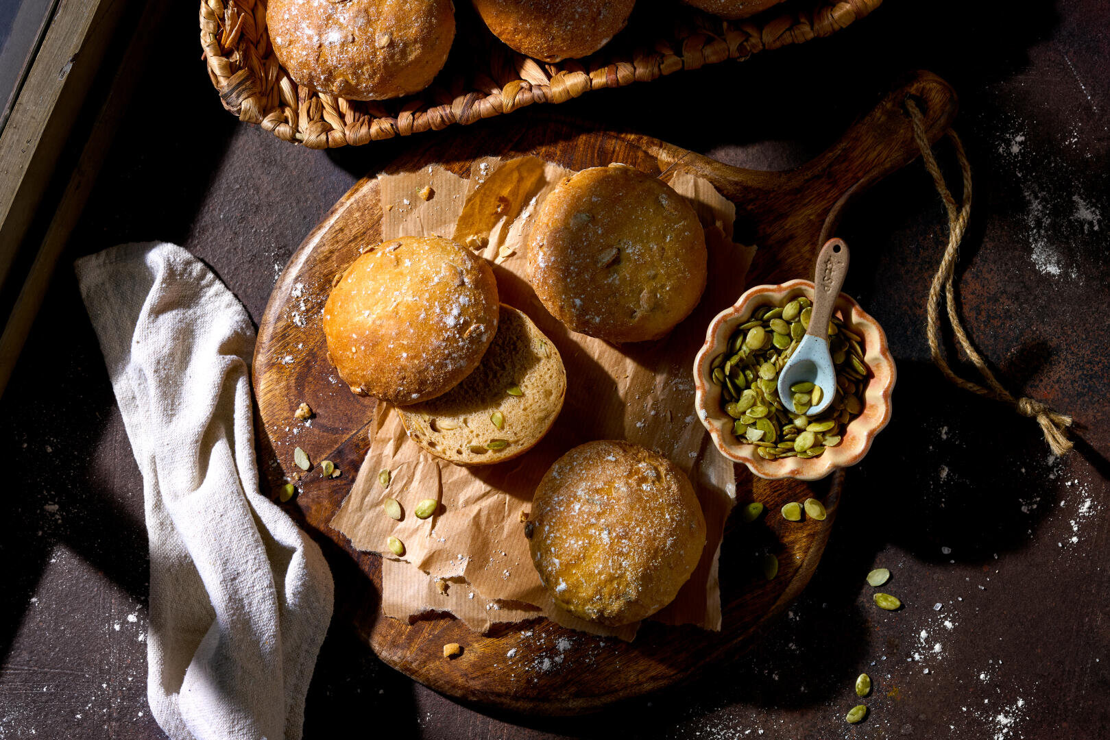 Runde Brötchen, bestreut mit Kürbiskernen und Puderzucker, liegen auf Backpapier auf einem Holzbrett. Daneben steht eine kleine Schale mit Kürbiskernen und einem Löffel. Daneben liegt ein leichtes Tuch.
