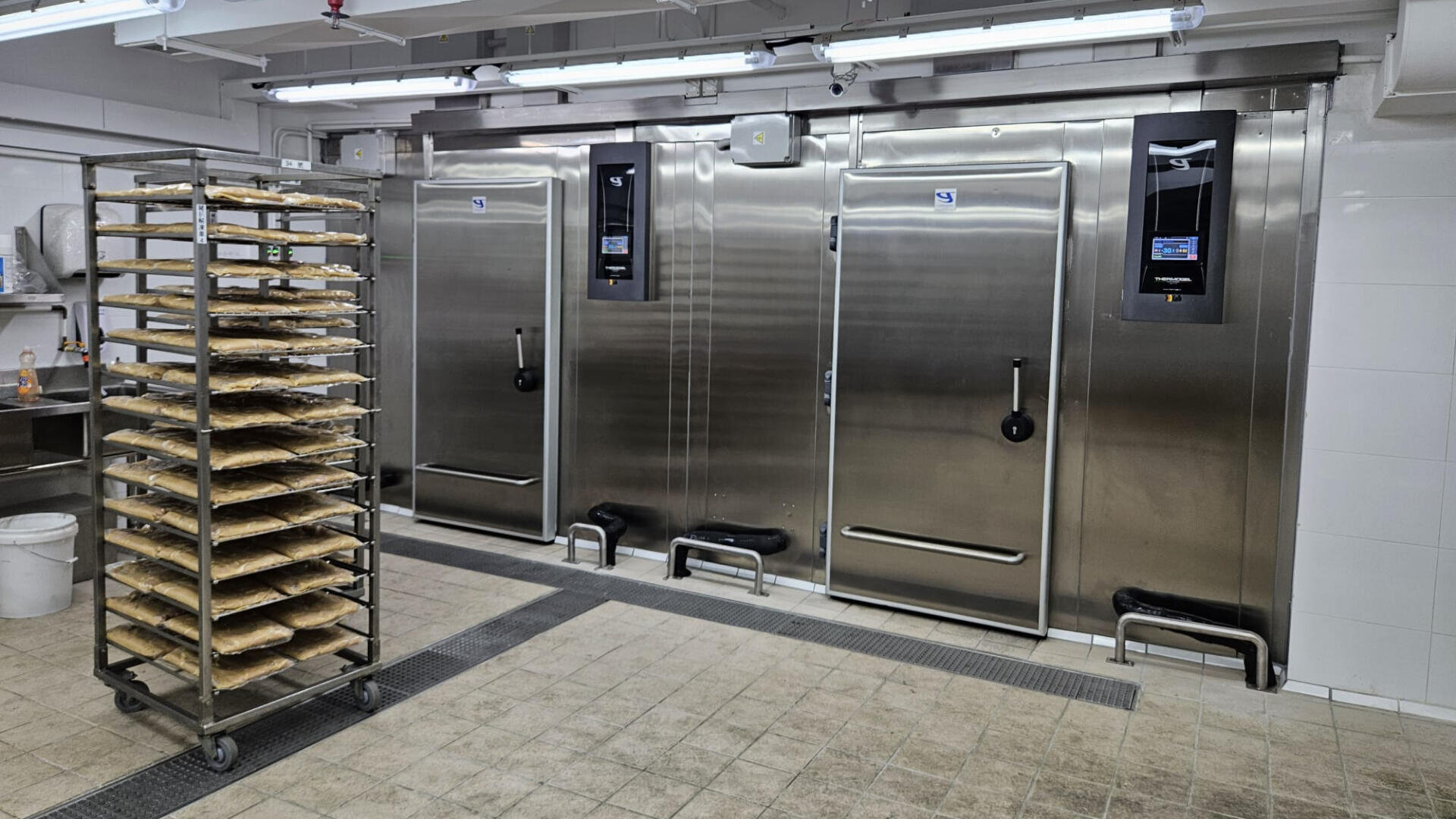A commercial kitchen with three large stainless steel industrial refrigerators and a rolling rack holding multiple trays of baked goods. The room has tiled floors, white walls, and bright overhead lighting.