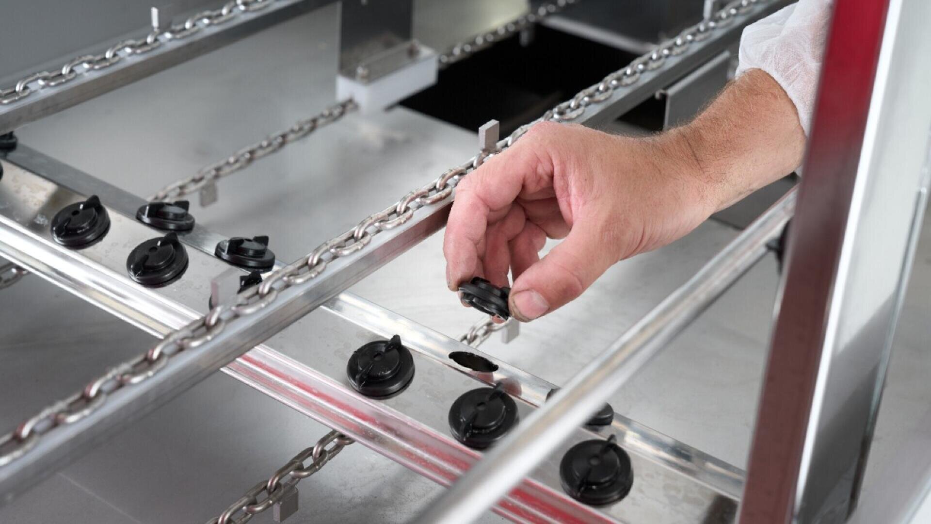 A person’s hand places a black plastic cap onto a metal component of an industrial machine, with a series of chains and cap-like objects aligned along metal rails.