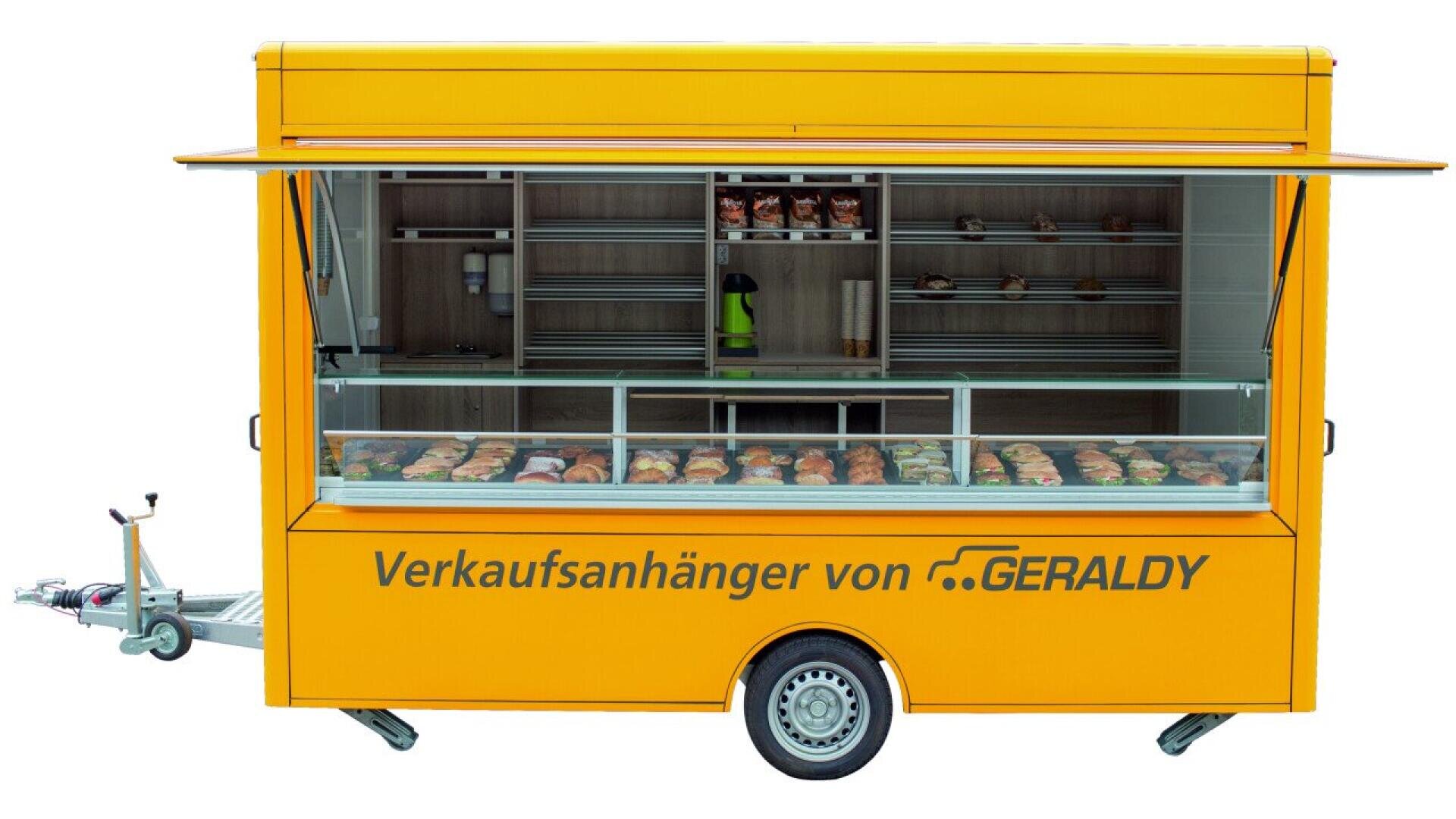 A yellow mobile food trailer with open service windows, displaying a variety of baked goods inside. The side reads “Verkaufsanhänger von GERALDY,” and shelves are stocked with bread and pastries.