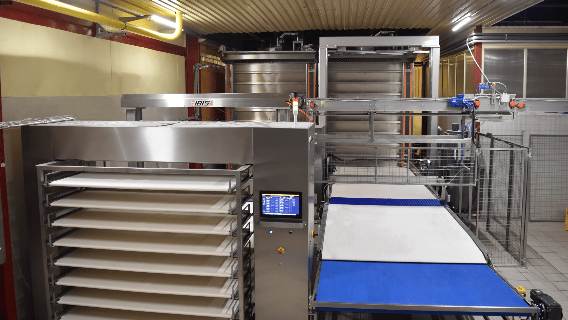 A commercial baking machine with stacked trays, a digital control panel, and conveyor belts in a brightly lit industrial kitchen setting.