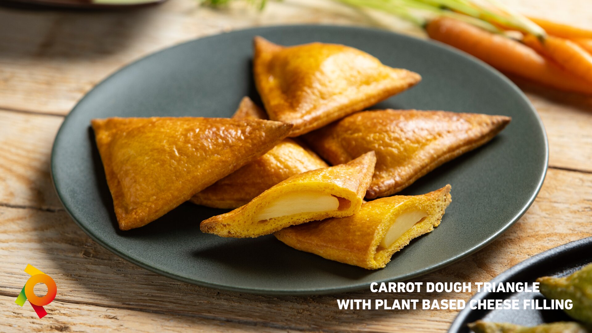 A plate with golden, triangular pastries, some cut open to show a plant-based cheese filling, sits on a wooden table. Text reads: Carrot dough triangle with plant based cheese filling.