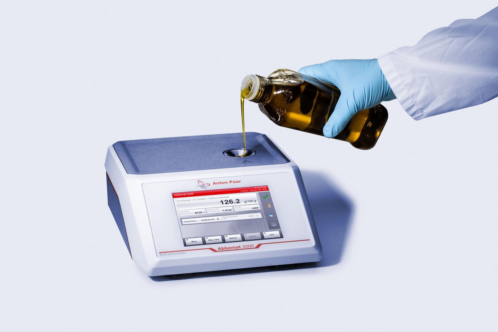 Eine Person, die einen blauen Handschuh trägt, gießt Olivenöl aus einer Flasche in ein Laborgerät mit einer digitalen Anzeige mit der Bezeichnung Abbe-Refraktometer Abbemat 3000.