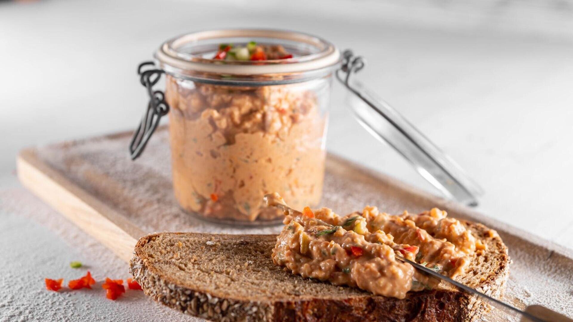 A slice of brown bread is spread with a chunky, creamy dip containing visible red and green vegetable pieces. Behind it, a glass jar filled with the same spread sits on a wooden board.