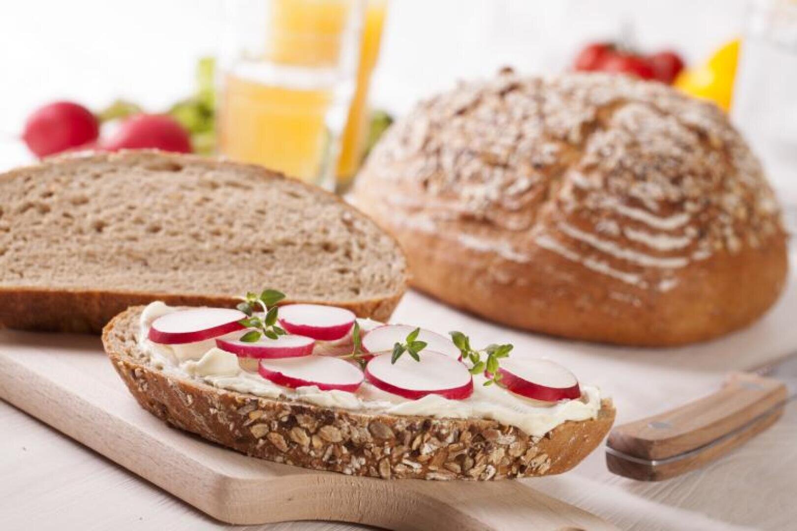 Eine Scheibe Vollkornbrot mit Frischkäse, Radieschenscheiben und frischen Kräutern liegt auf einem Schneidebrett, neben einem Laib Brot und einem Glas Orangensaft.