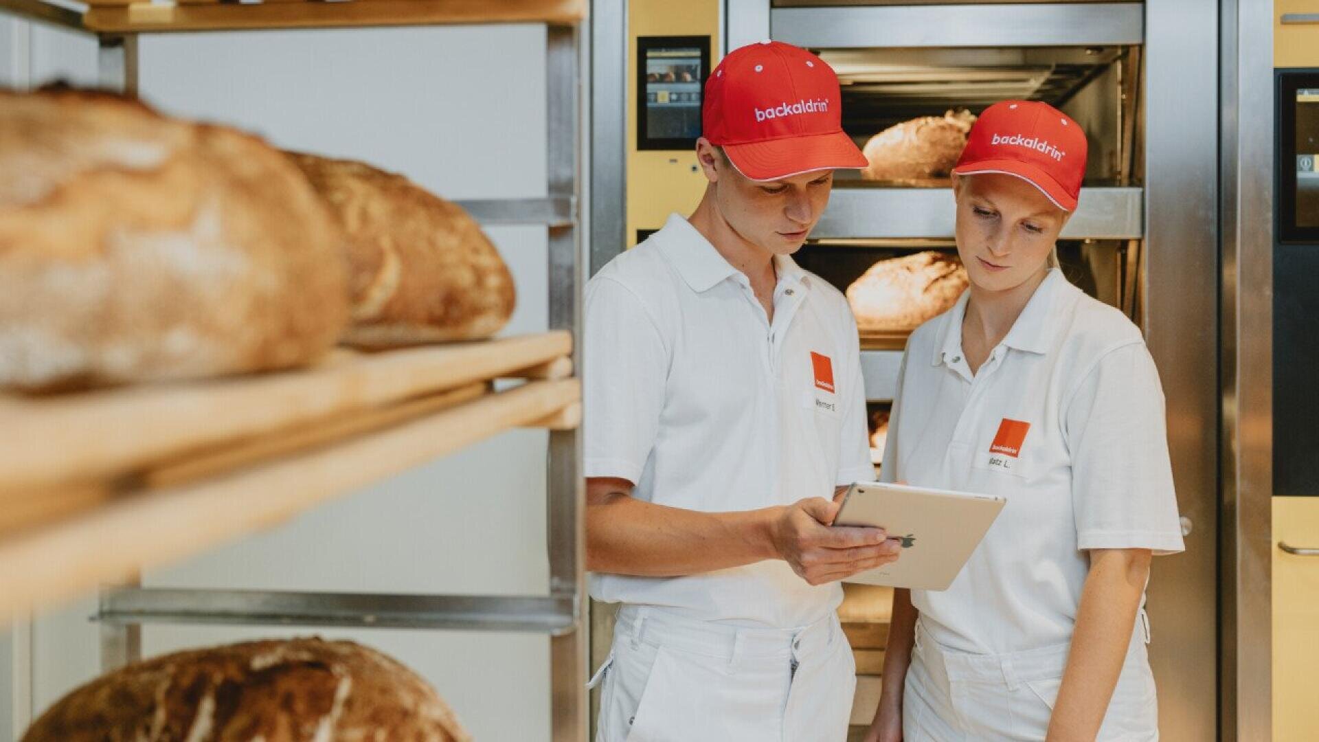 Two bakery workers in white uniforms and red hats stand by shelves of bread, looking at a tablet together in a modern bakery setting.