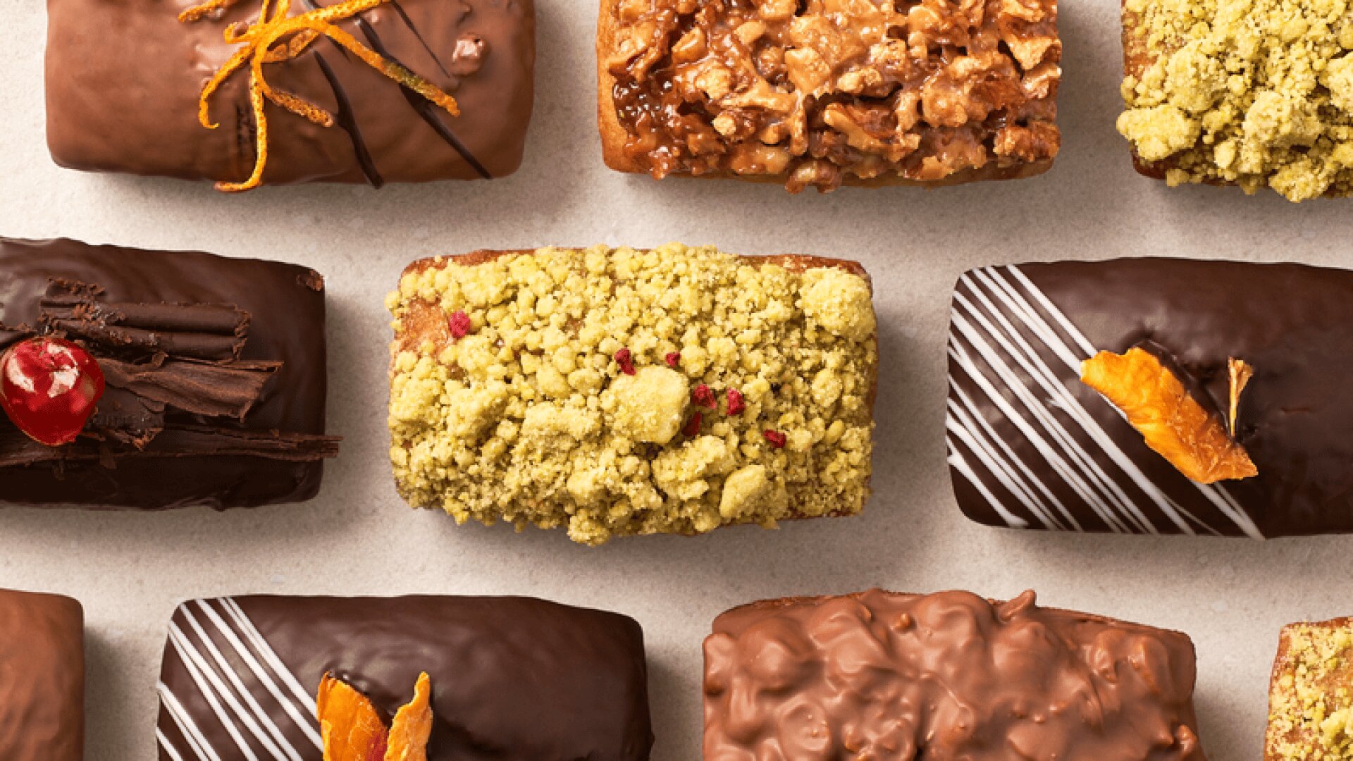 An assortment of rectangular chocolate and nut-covered pastries, some topped with candied fruit, orange zest, or pistachio crumbs, arranged in neat rows on a light surface.