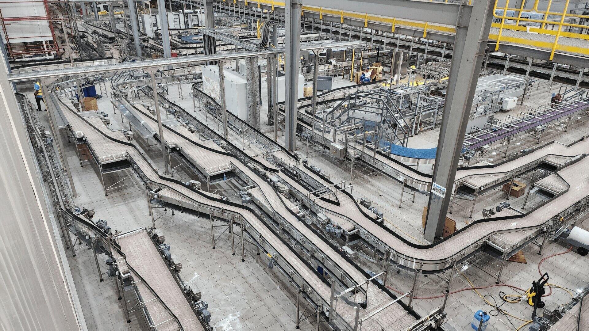 A large, modern factory with multiple empty conveyor belts winding through an industrial space. A few workers in yellow helmets are seen on the shiny tiled floor among the machinery.