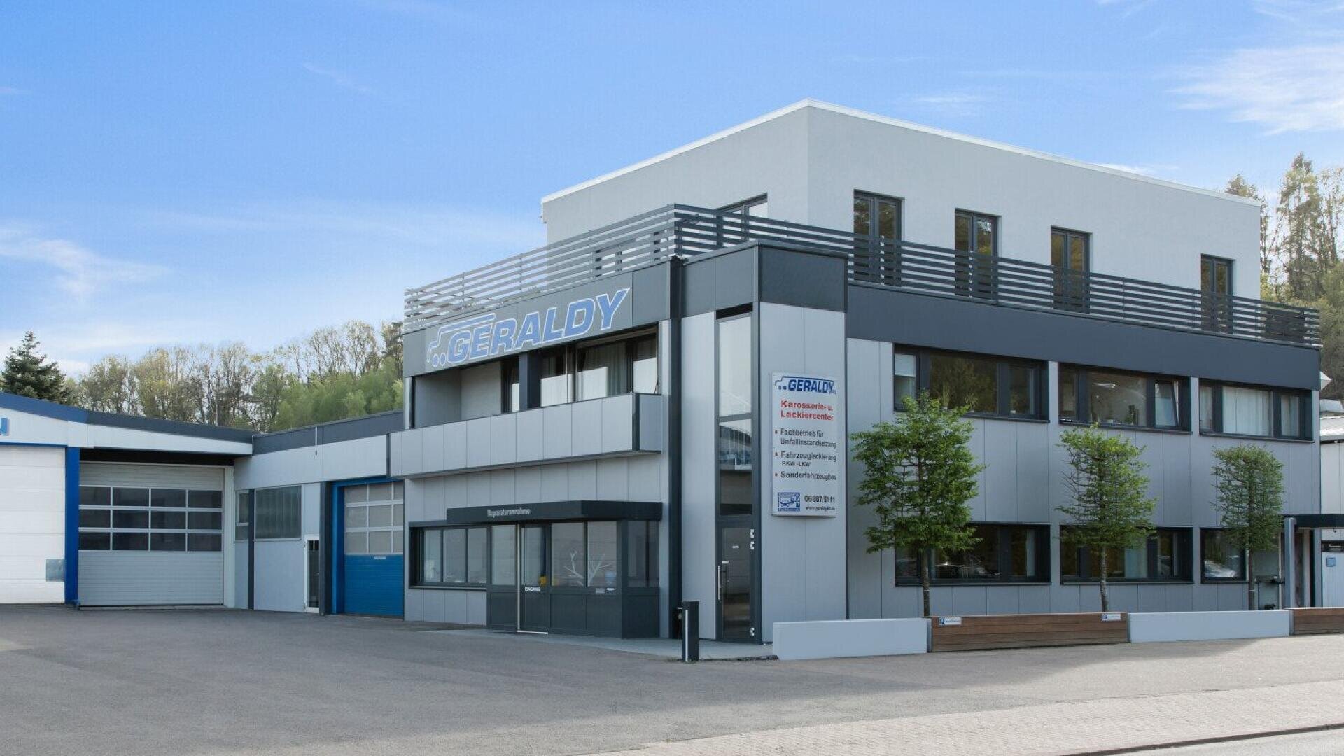 A modern, two-story commercial building with large windows, gray and white exterior panels, and GERALDY signage on the facade. Small trees line the front, and the area is clean and paved.