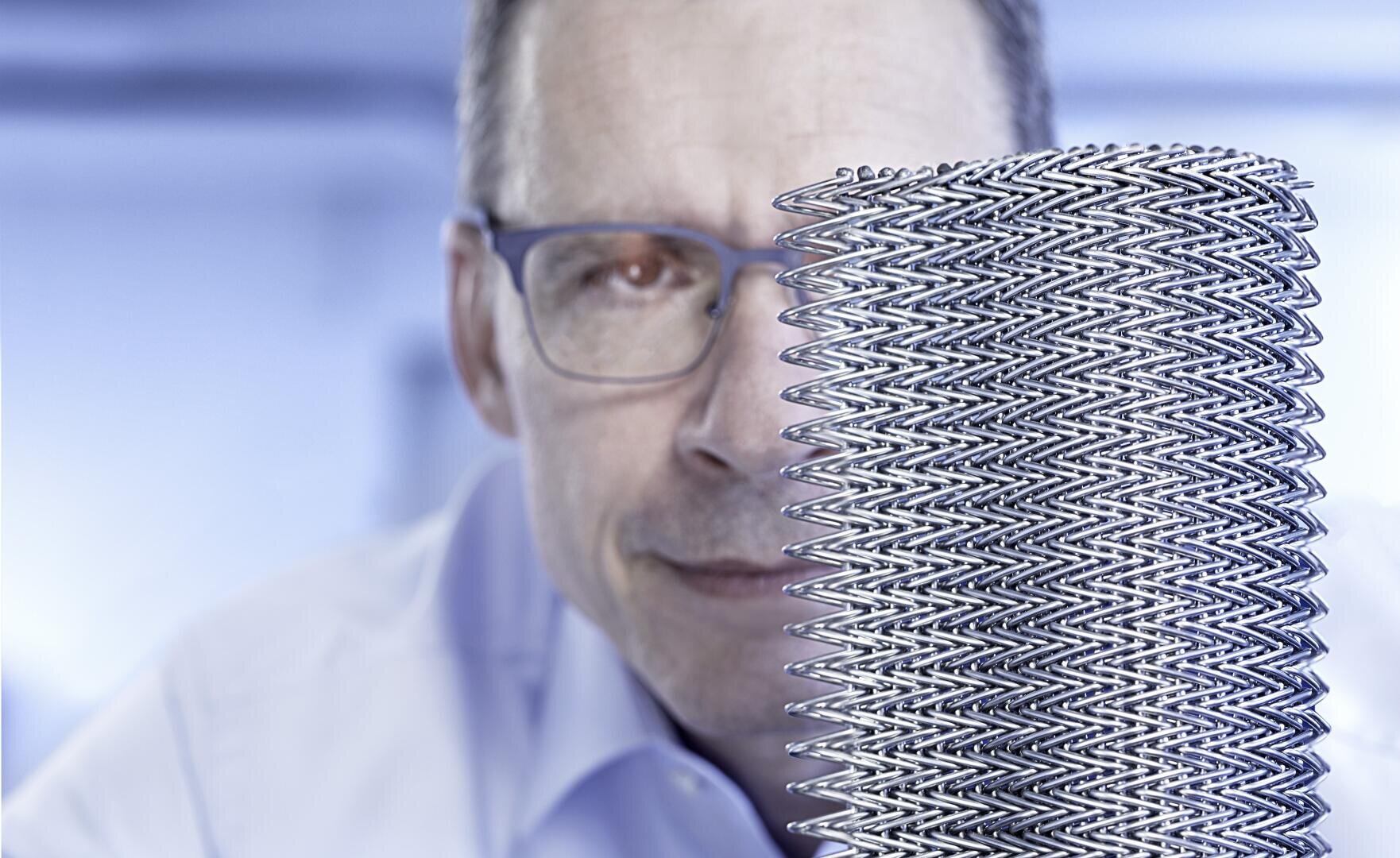 A man with glasses looks at a large, cylindrical metal mesh that can be seen in the foreground, with a blue-tinted, blurred background.