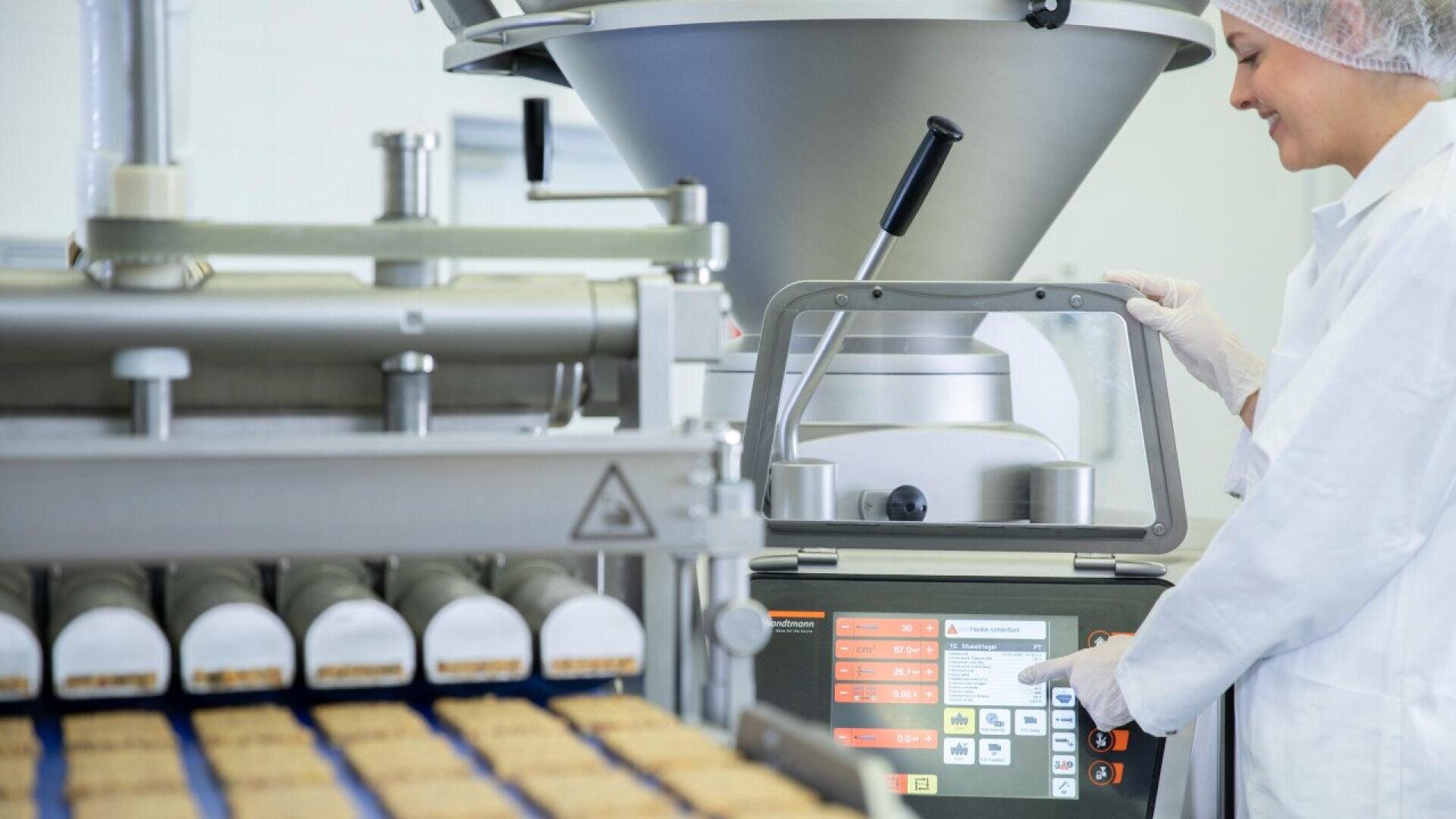 A person in a lab coat, hair net, and gloves operates a touchscreen control panel on a large industrial food processing machine producing rows of rectangular food items on a conveyor belt.
